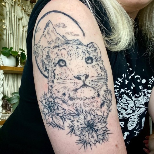 Snow leopard with mountains and flowers realism tattoo on the upper arm by Thylacine Tattoo & Art, Los Angeles, California