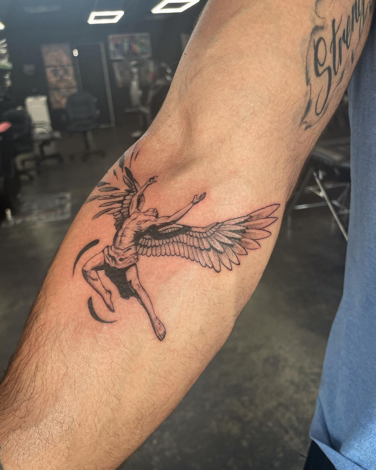 Winged creature carrying a woman black and grey tattoo on the forearm by Tattoos by Tessa