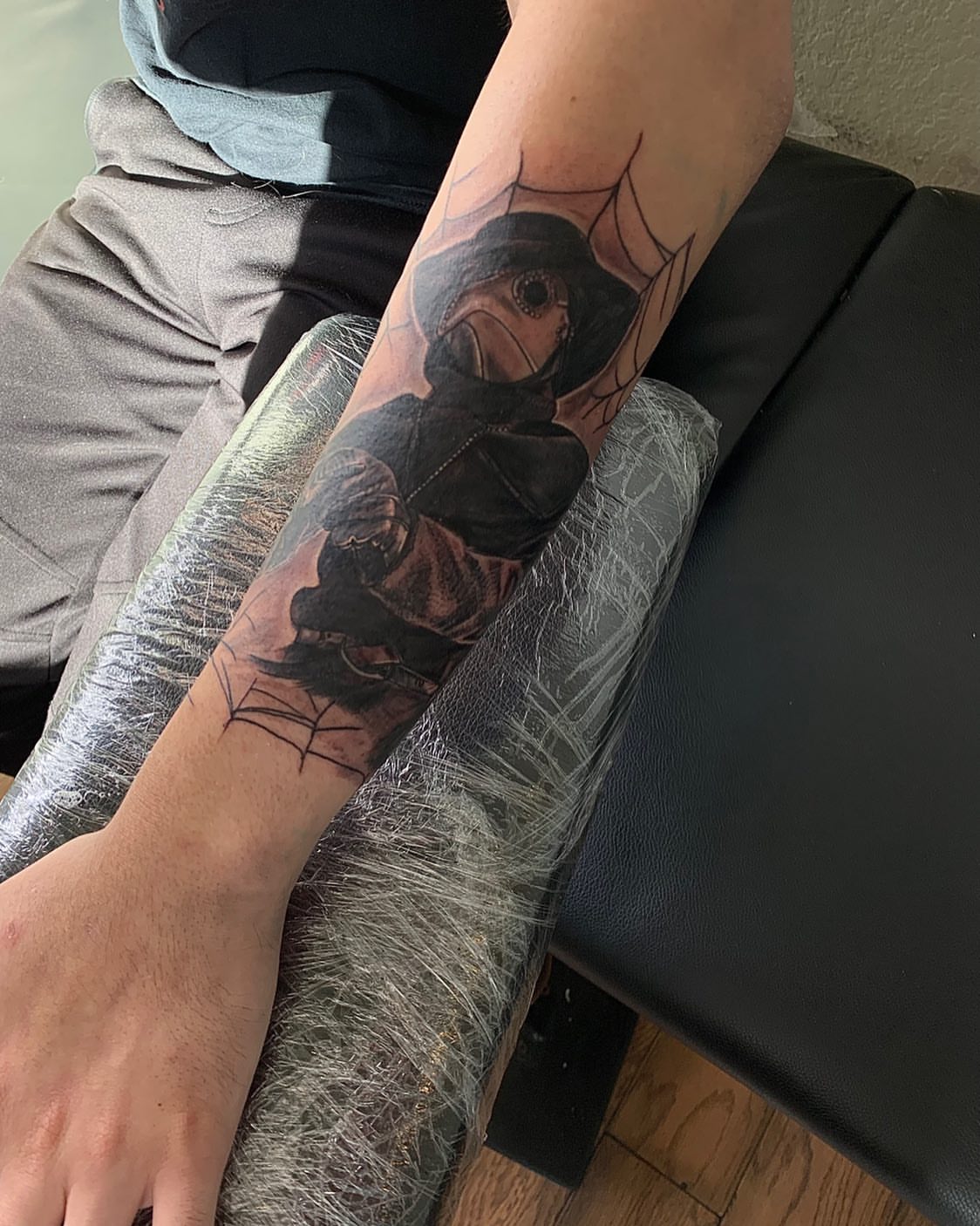 Mask-wearing figure black and grey tattoo on the forearm by Leo Torres