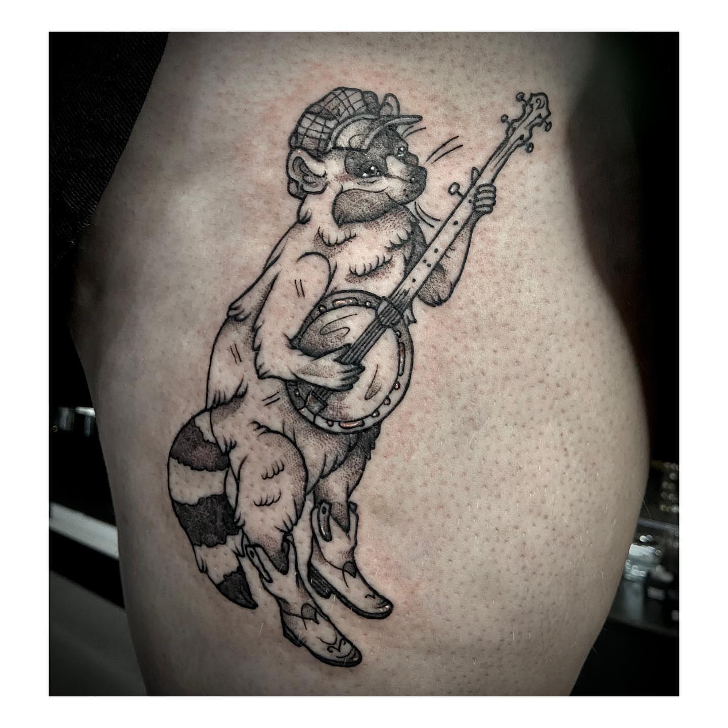 Raccoon playing banjo black and grey tattoo on the thigh by Luna
