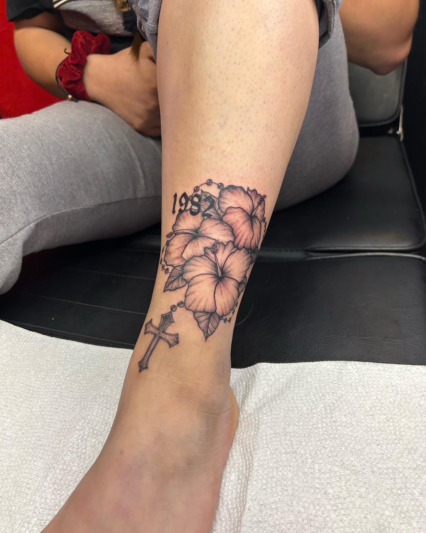 Cross and hibiscus flowers black and grey tattoo on the forearm by Tattoo Artist