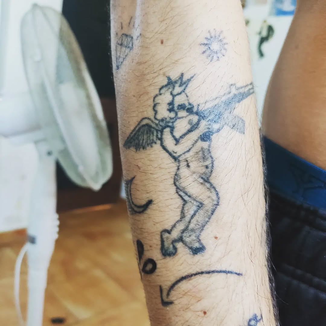 Cherub with a gun minimalist tattoo on the forearm by Tatuadora & Piercer