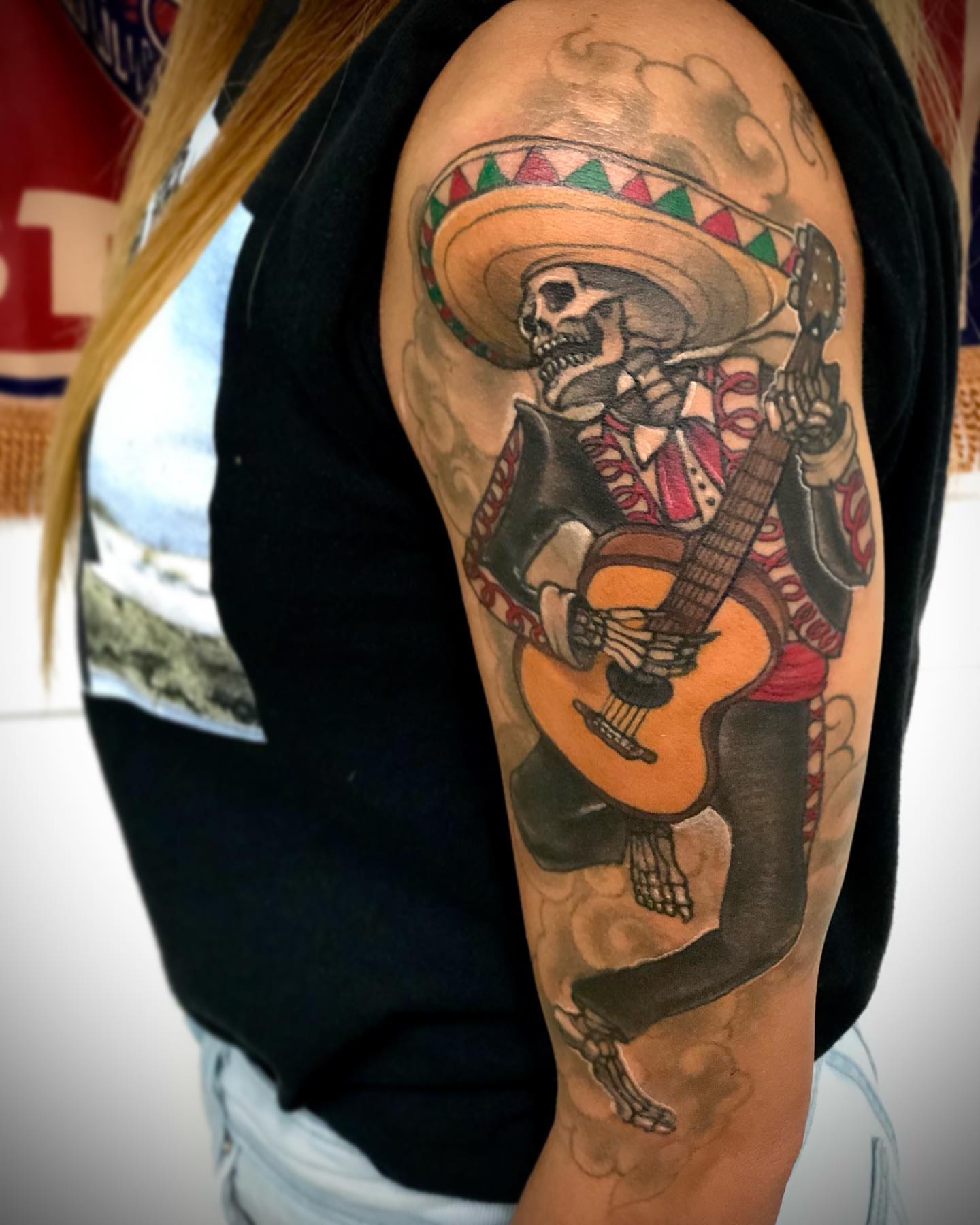Skeleton playing guitar Chicano tattoo on the upper arm by Alec Bauer