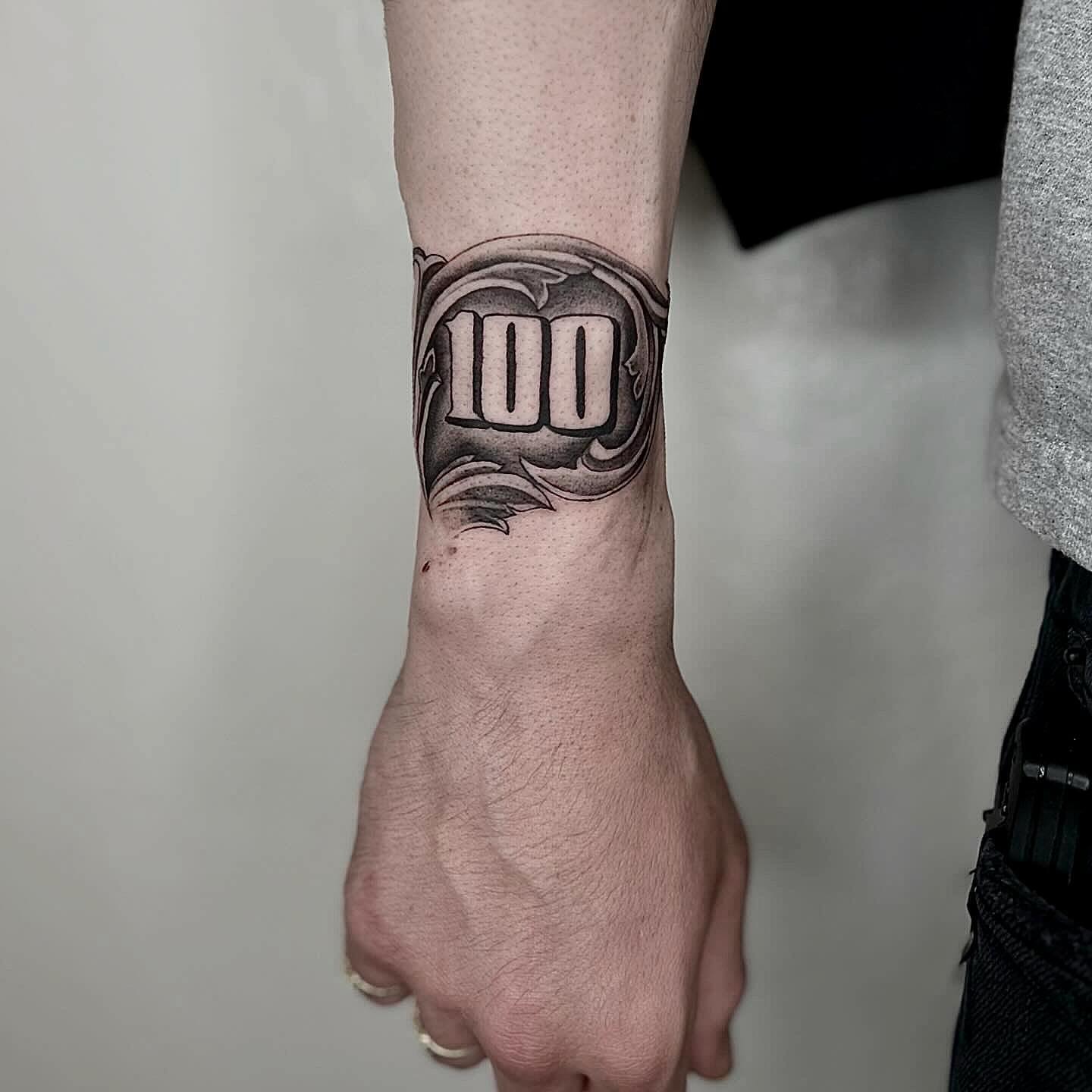 Number 100 black and grey tattoo on the forearm by Jocelyn