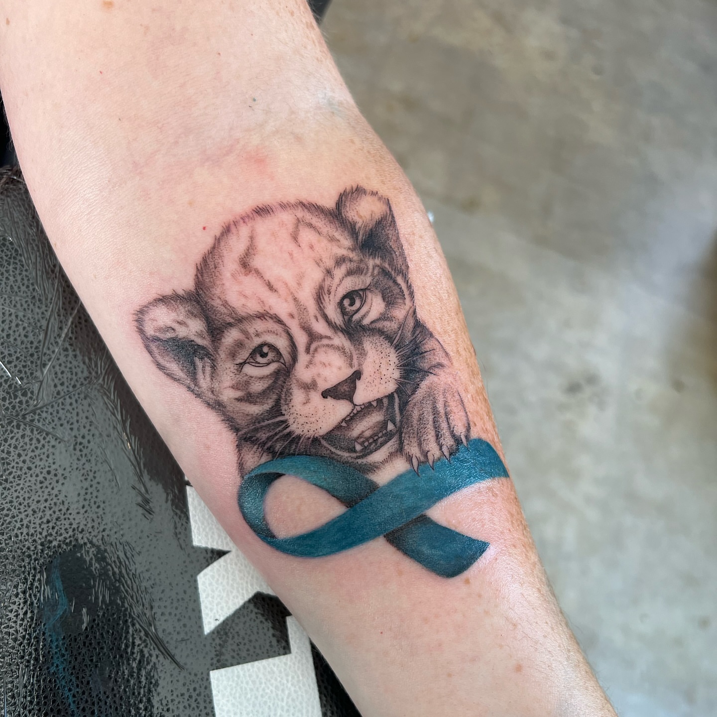 Lion cub with teal ribbon realism tattoo on the forearm by Dom