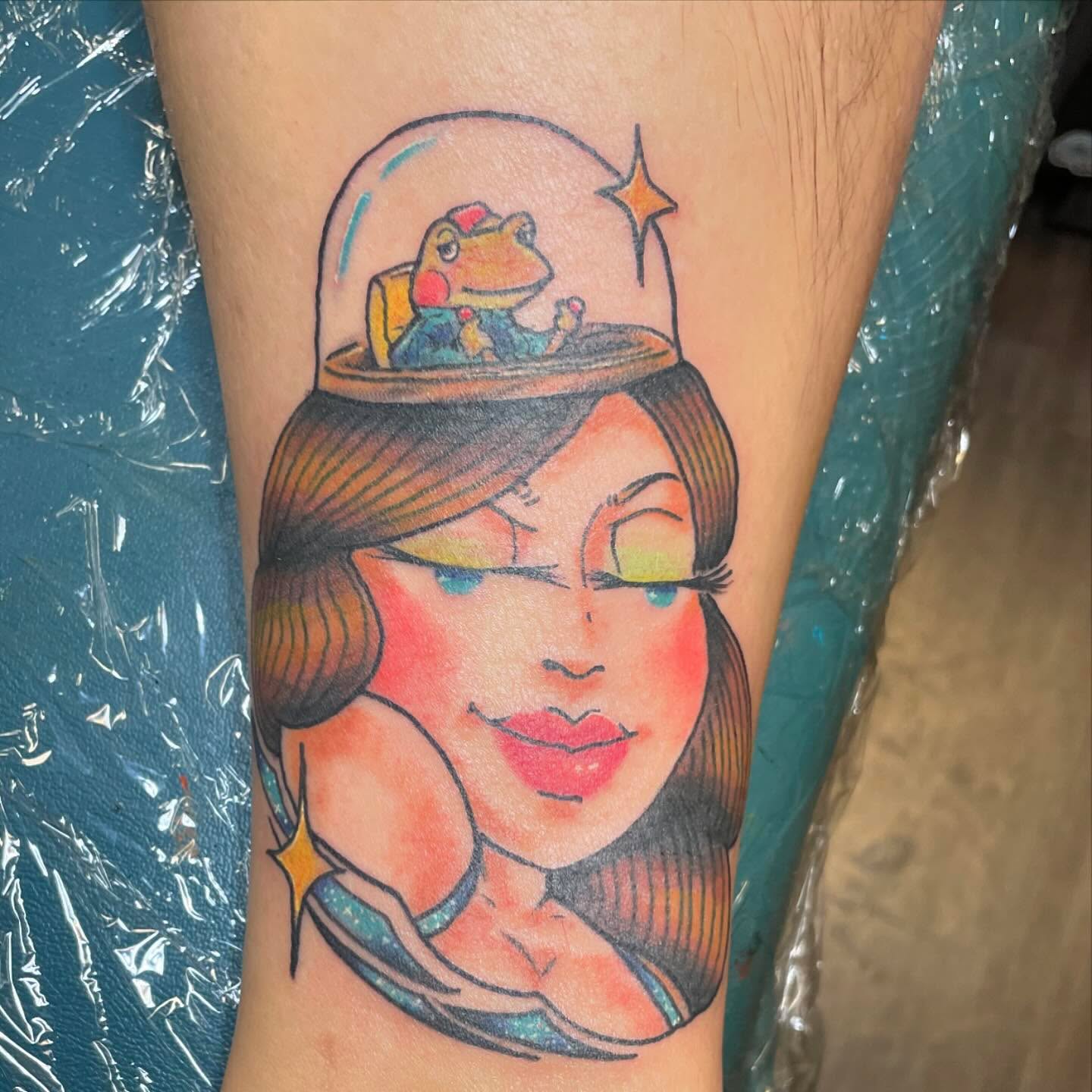 Woman with frog in helmet illustrative tattoo on the forearm by Evan
