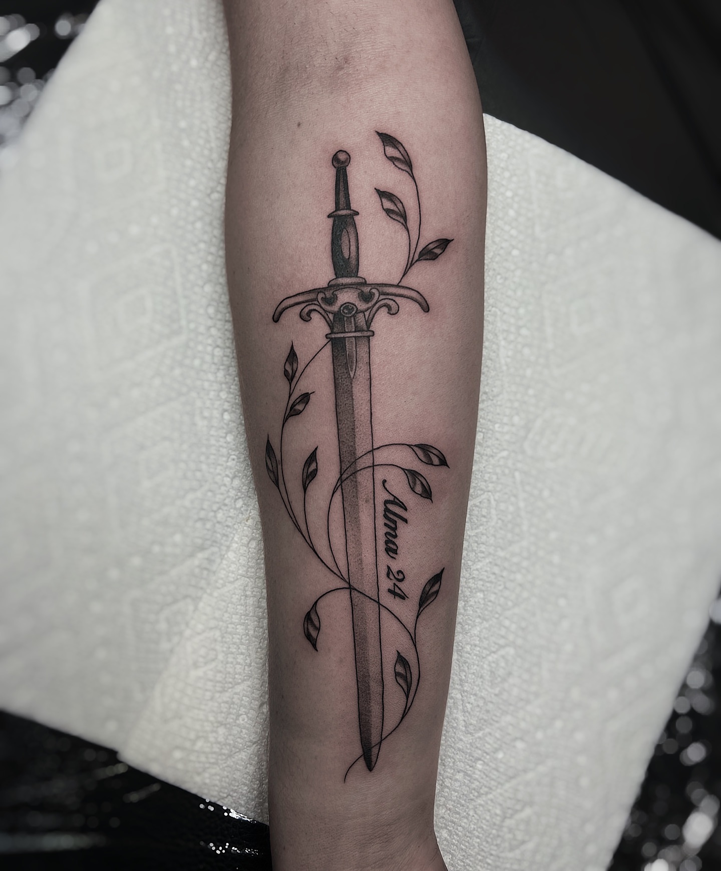 Sword and leaves black and grey tattoo on the forearm by Kolby
