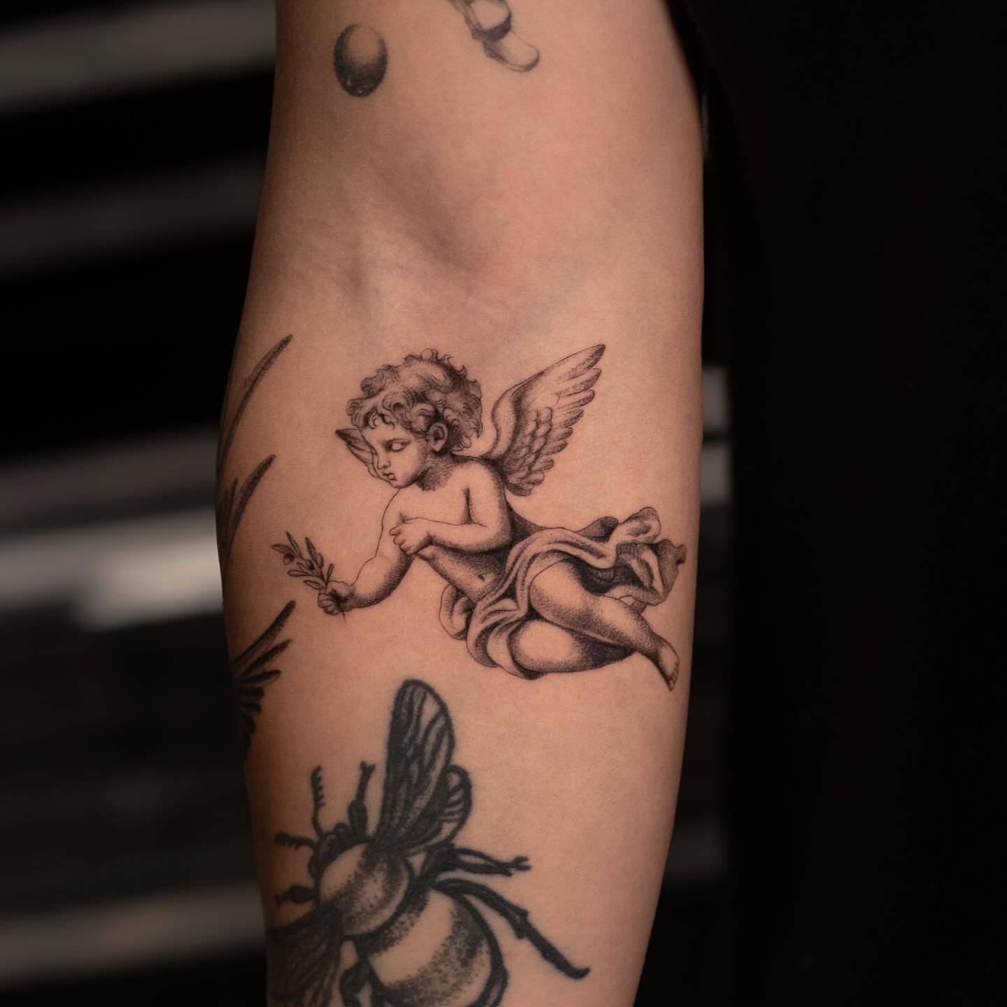 Cherub and bee black and grey tattoo on the forearm by Valery Cardona