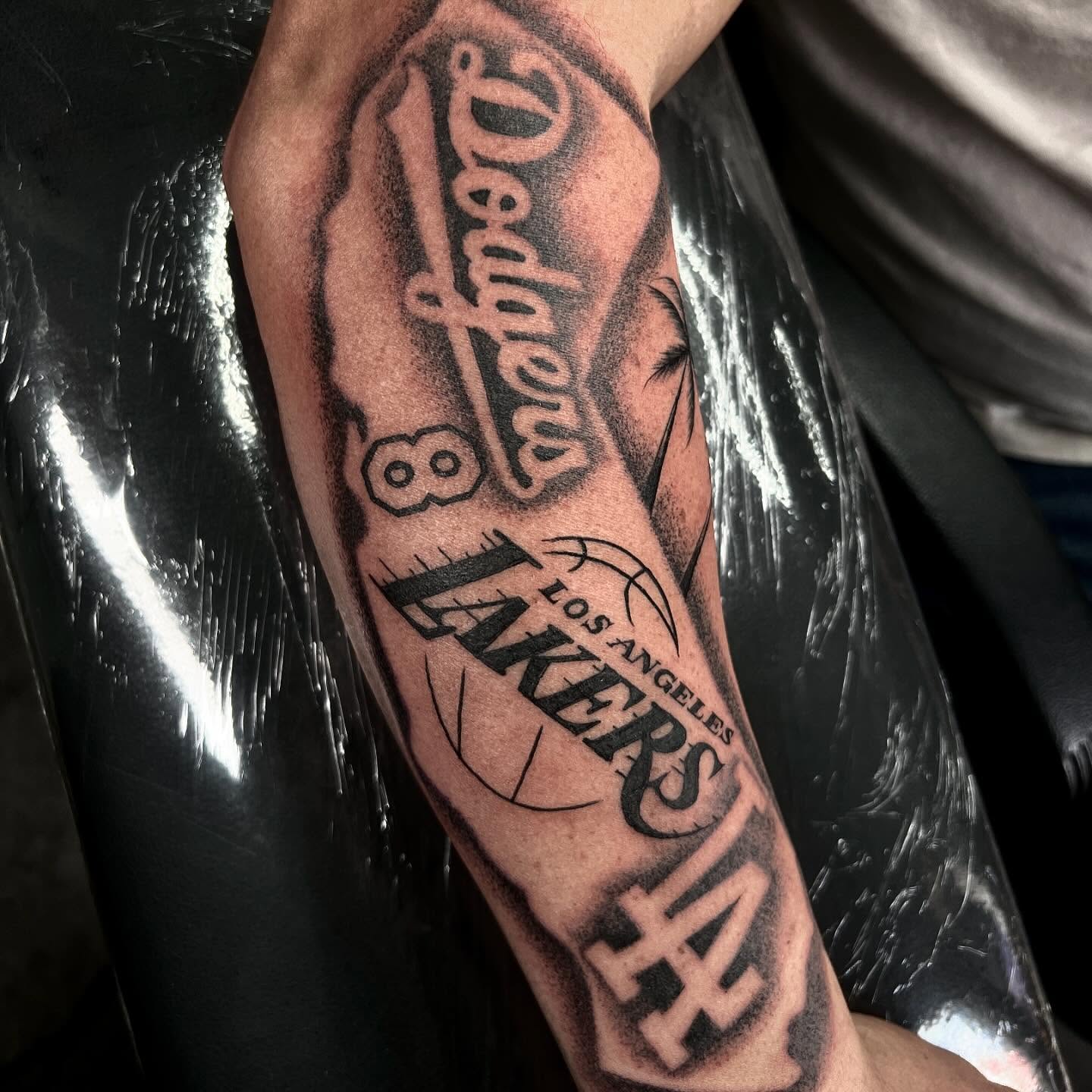 Dodgers and Lakers logos black and grey tattoo on the forearm by Joey Moreno