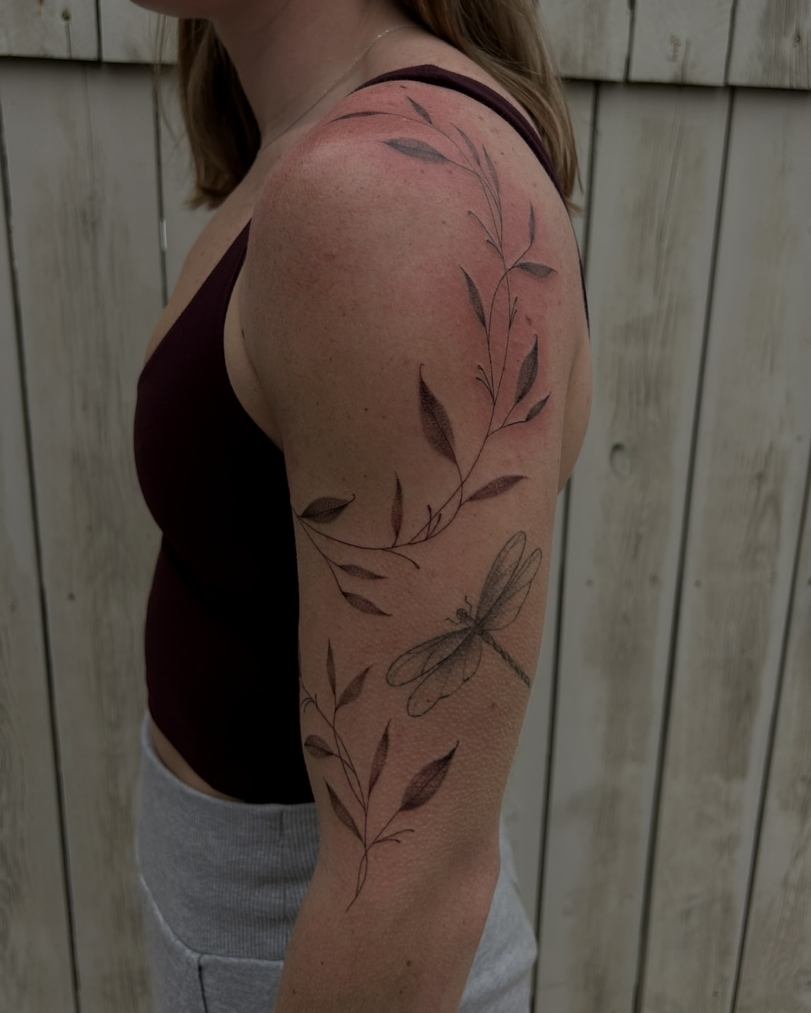 Dragonfly and leaves black and grey tattoo on the upper arm by Kennedy Ramey