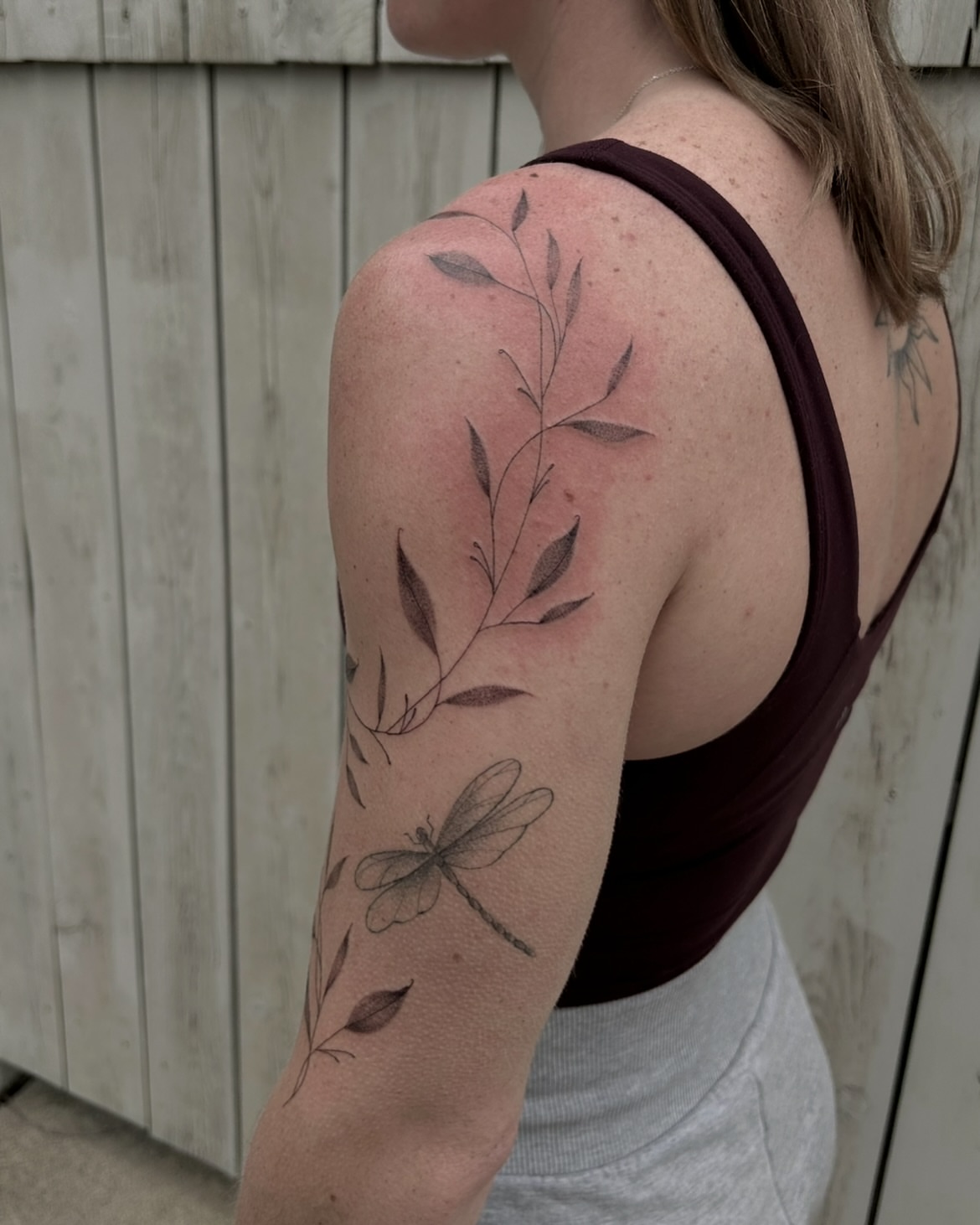Plant and dragonfly black and grey tattoo on the upper arm by Kennedy Ramey