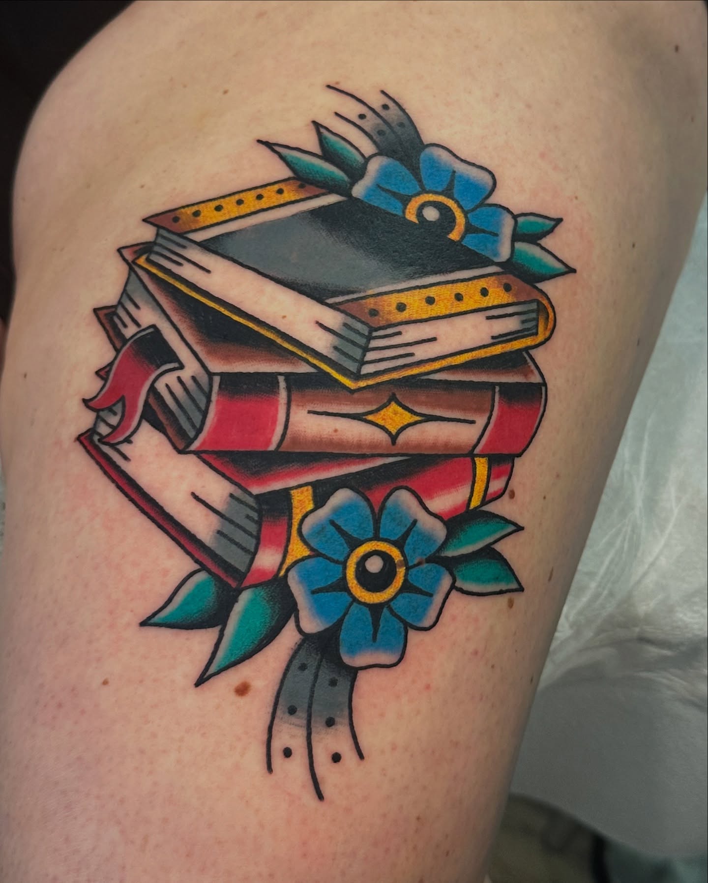 Stacked books and flowers American traditional tattoo on the thigh by Emily Masa