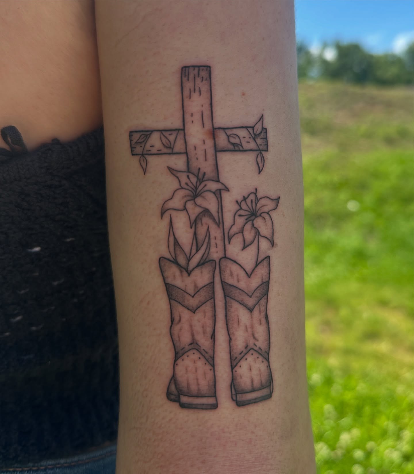 Cross with boots and lilies black and grey tattoo on the forearm by Imani