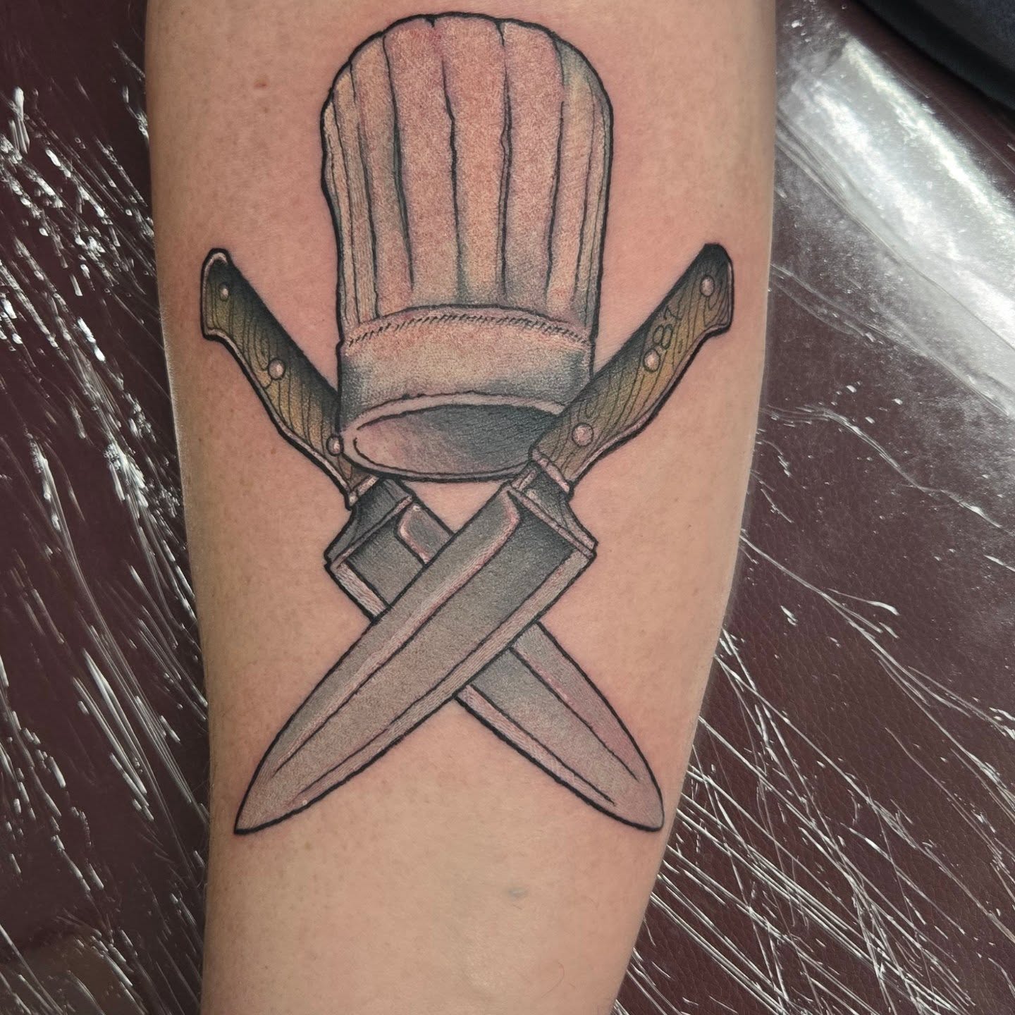 Chef hat and crossed knives black and grey tattoo on the forearm by Jamie Clinton