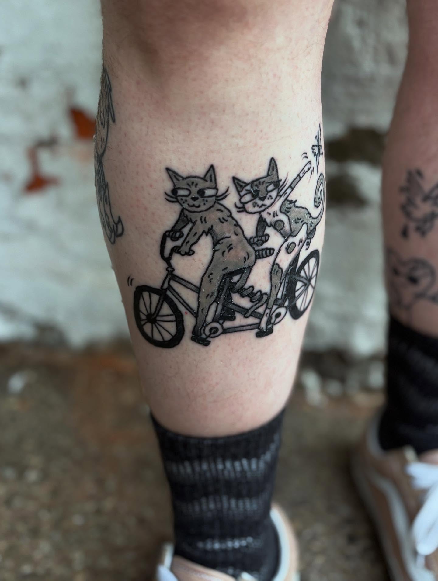 Cats riding tandem bicycle black and grey tattoo on the leg by kris noble floyd