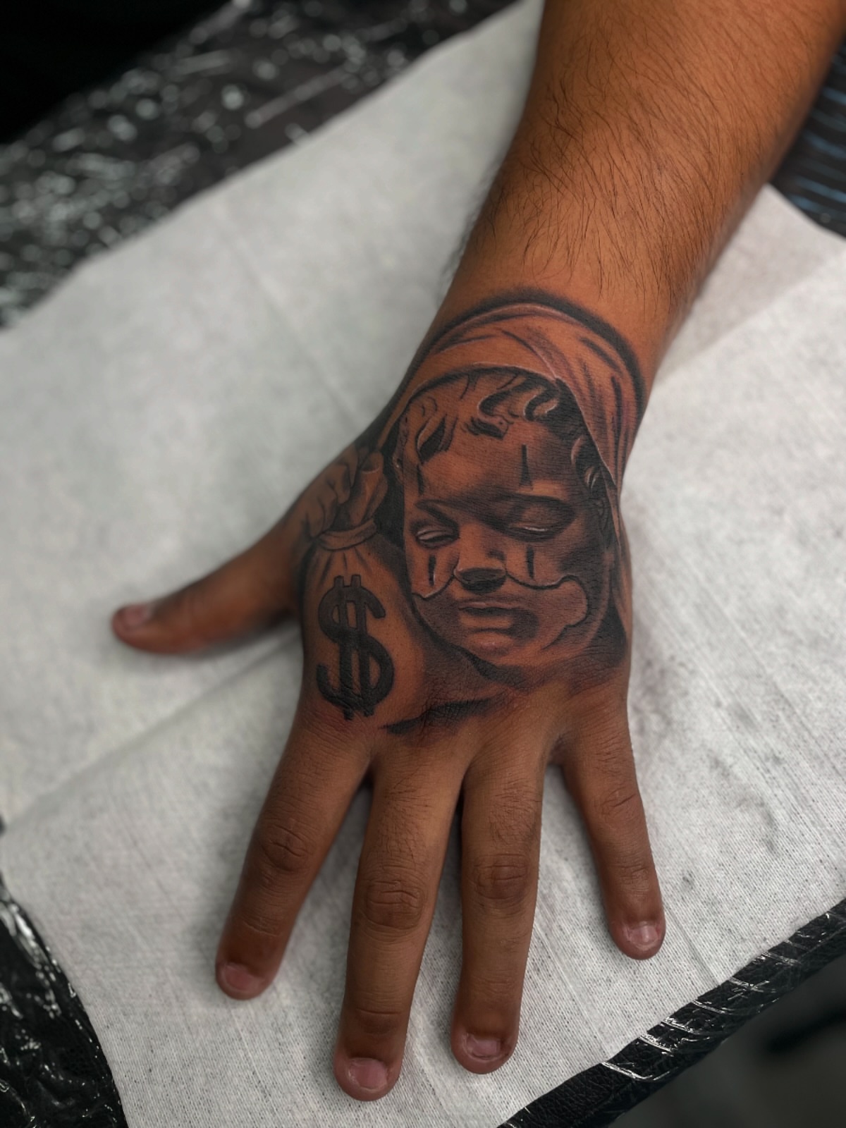 Mask with dollar sign black and grey tattoo on the hand by Vic