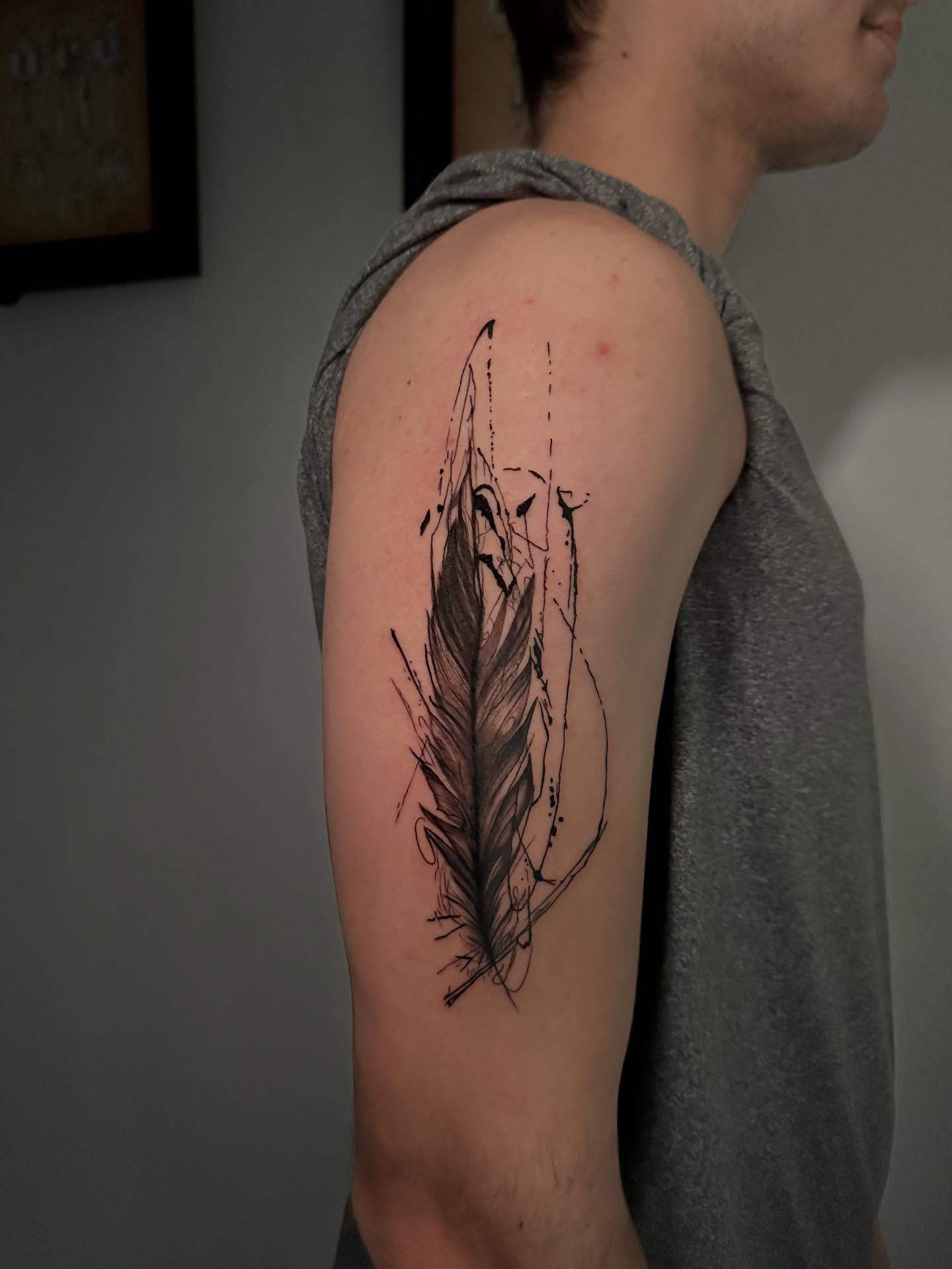 Feather black and grey tattoo on the upper arm by Élexus Foster