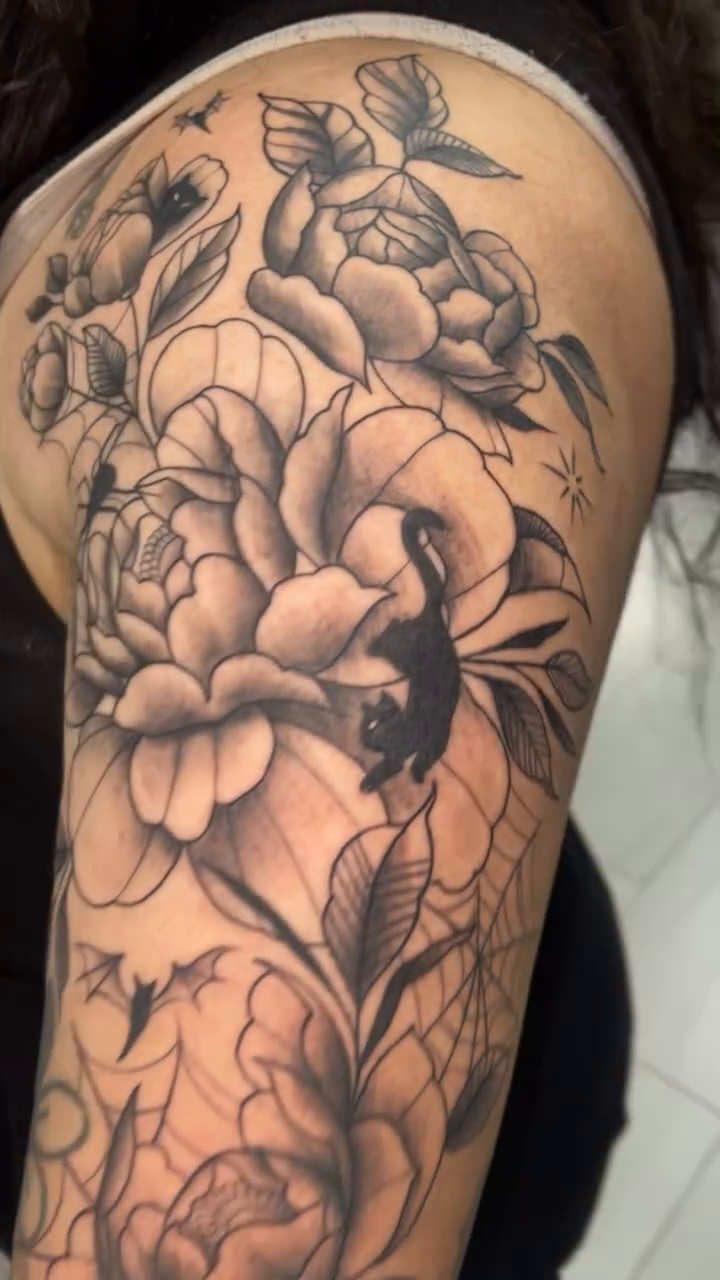 Peonies, spiders, and a black cat dotwork tattoo on the upper arm by Natalie