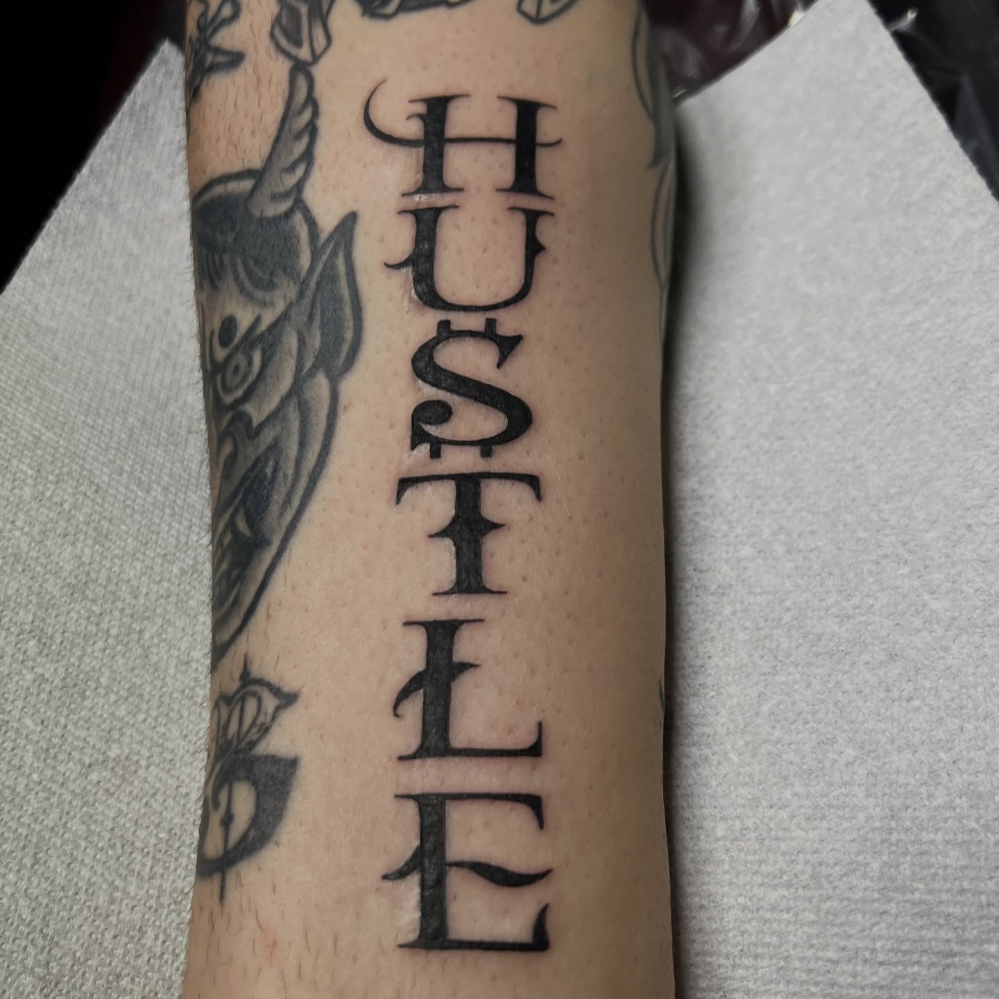 HUSTLE lettering script tattoo on the forearm by Nick Ochoa