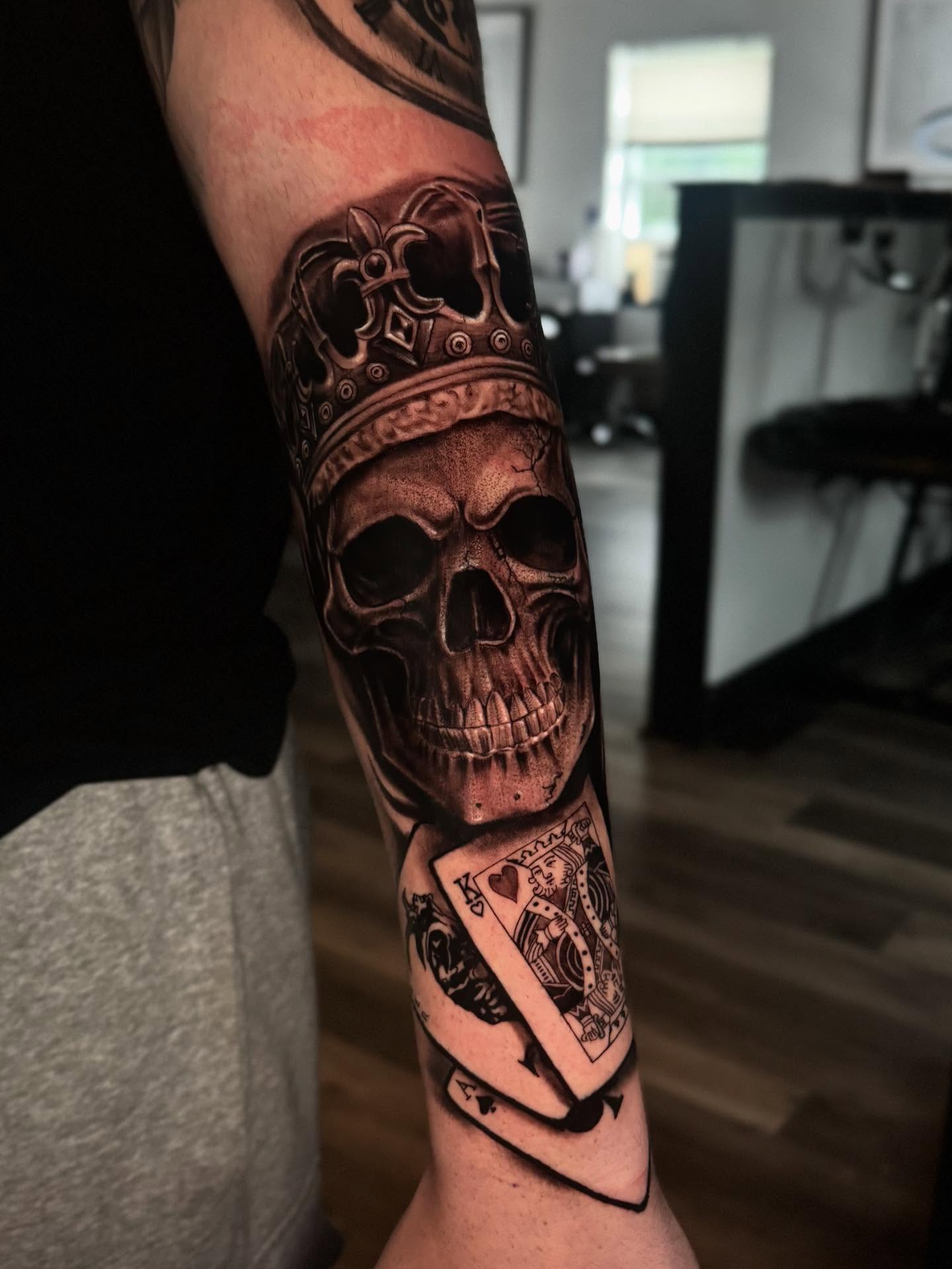 Skull with crown and playing cards realism tattoo on the forearm by Miguel Garcia