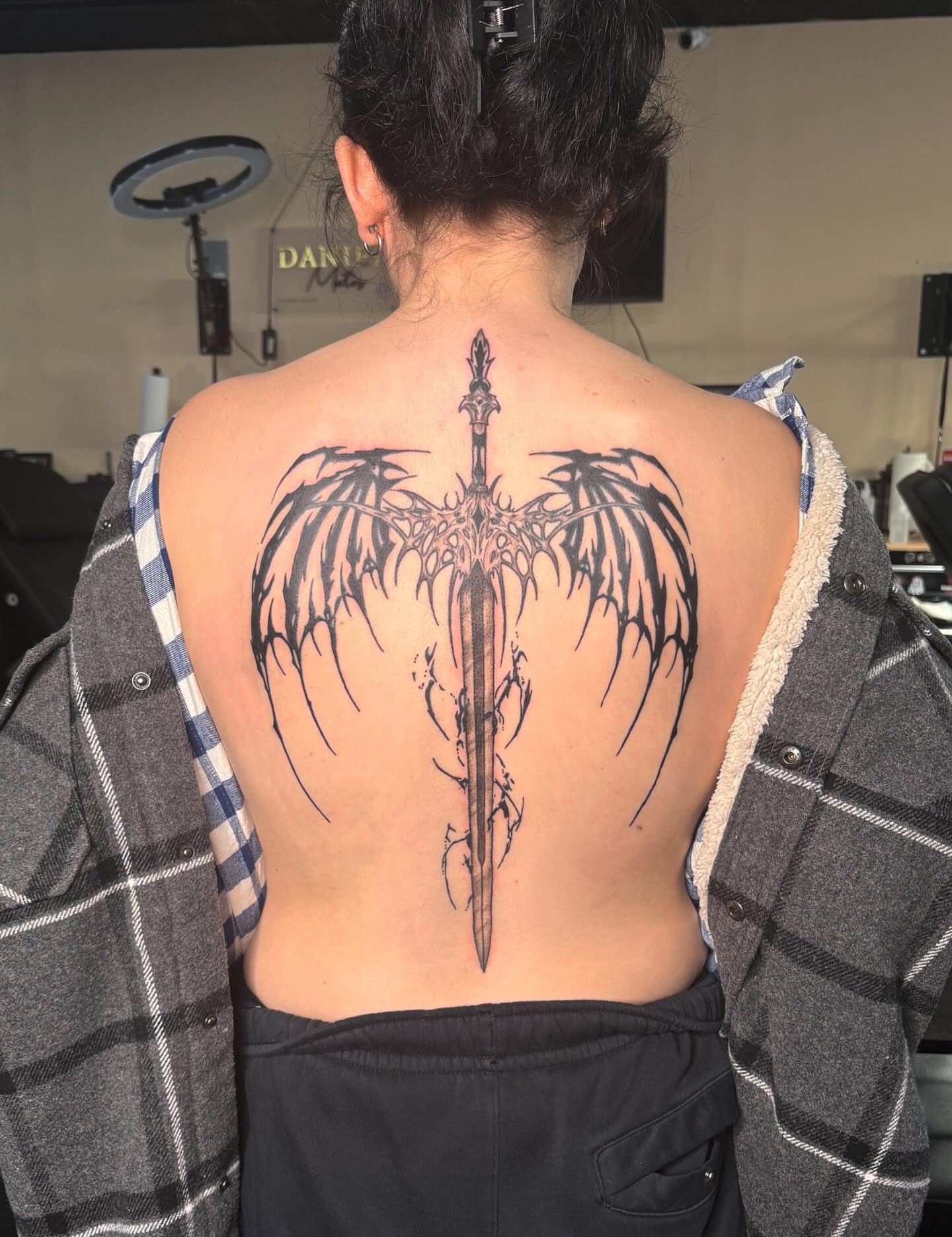 Sword with wings black and grey tattoo on the back by Tessa Simmons - Tattoo Artist