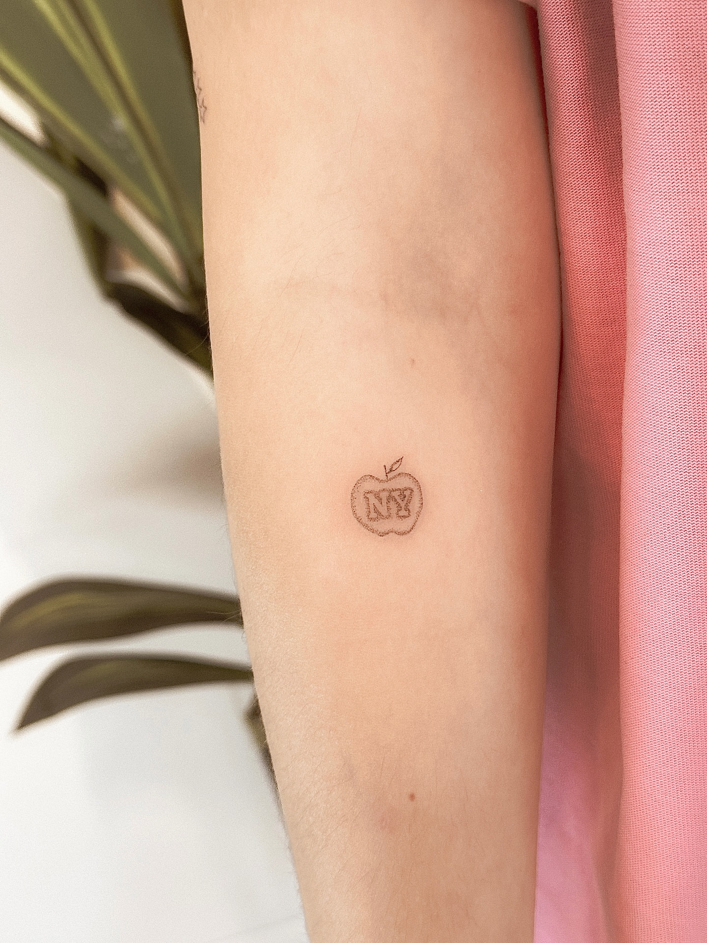Apple with 'NY' lettering script tattoo on the forearm by Yuli Noama