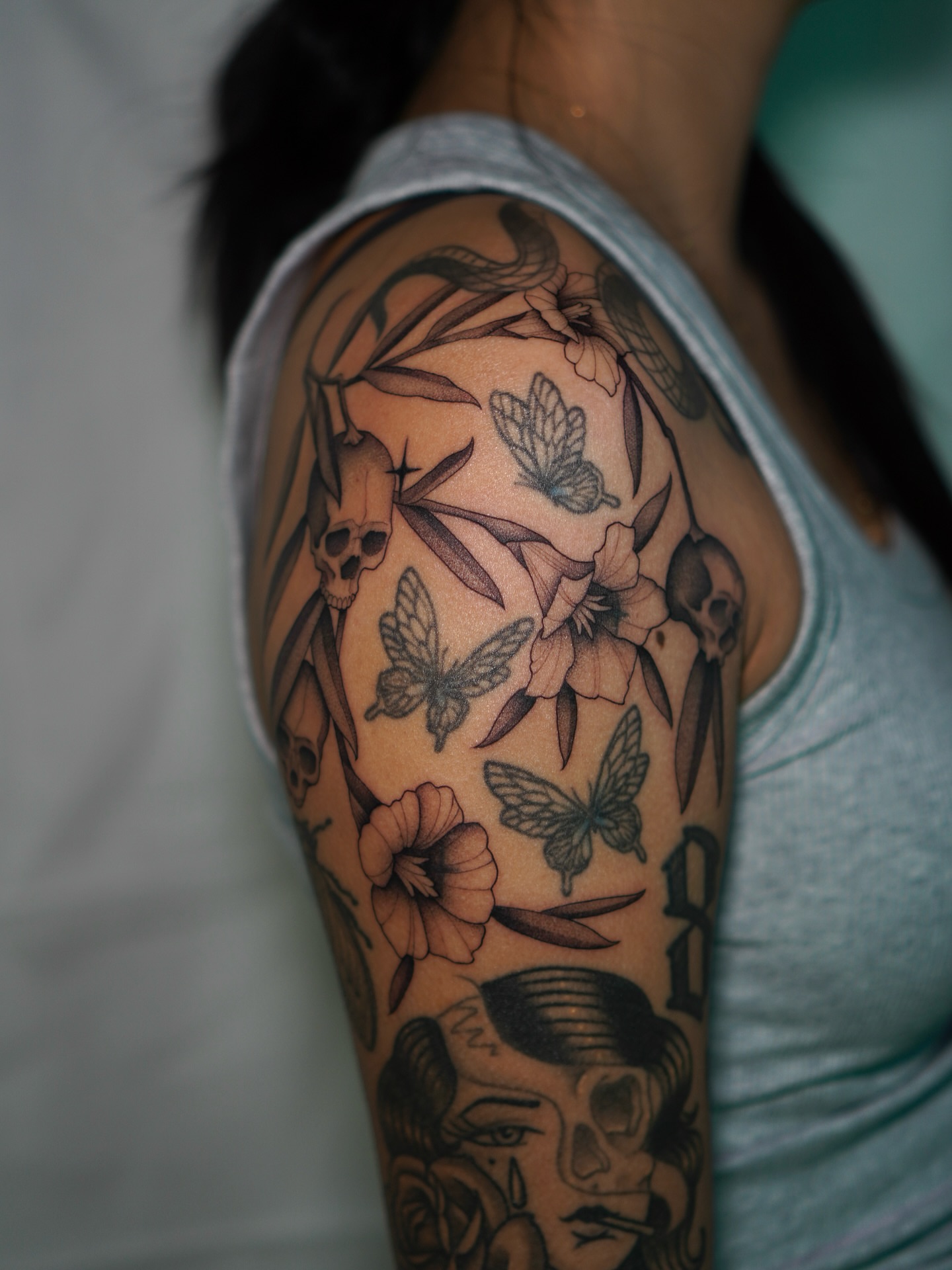 Skulls, butterflies, flowers black and grey tattoo on the upper arm by Carlos Abad