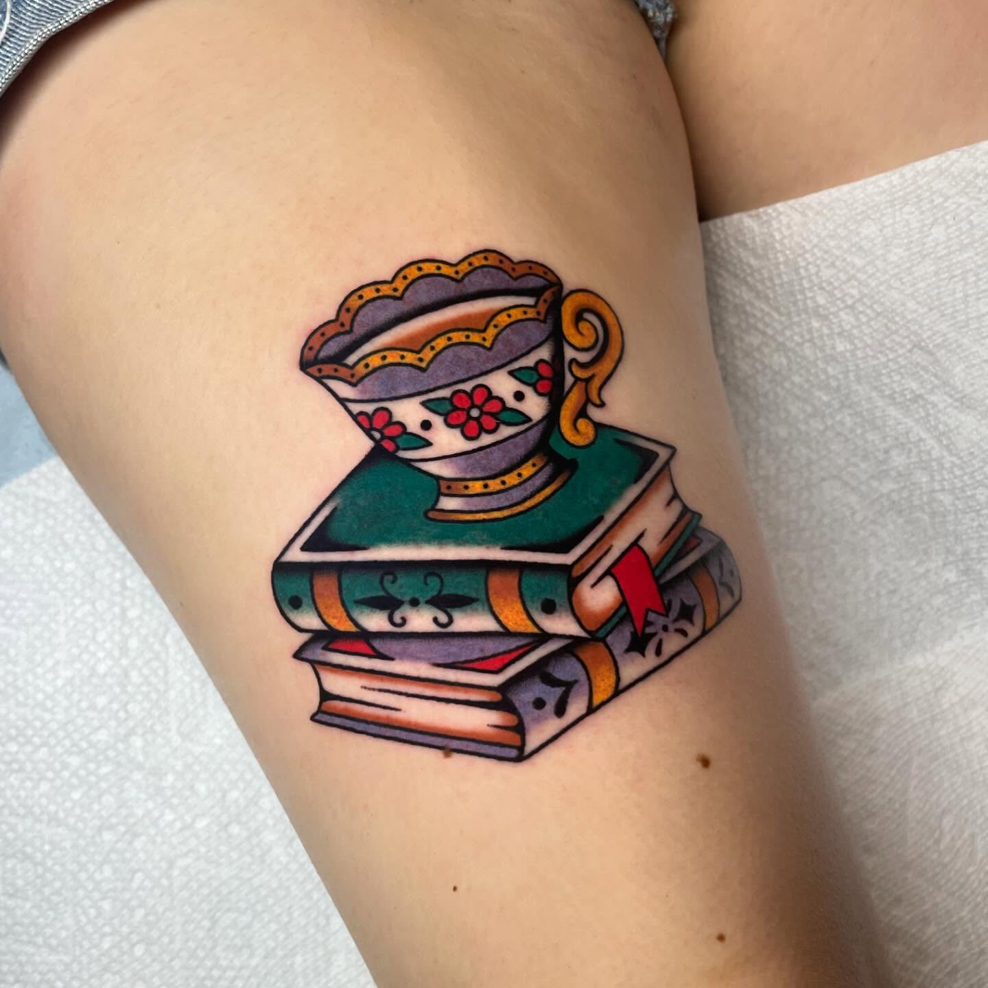 Stack of books with teacup American traditional tattoo on the thigh by Nova Vaughn