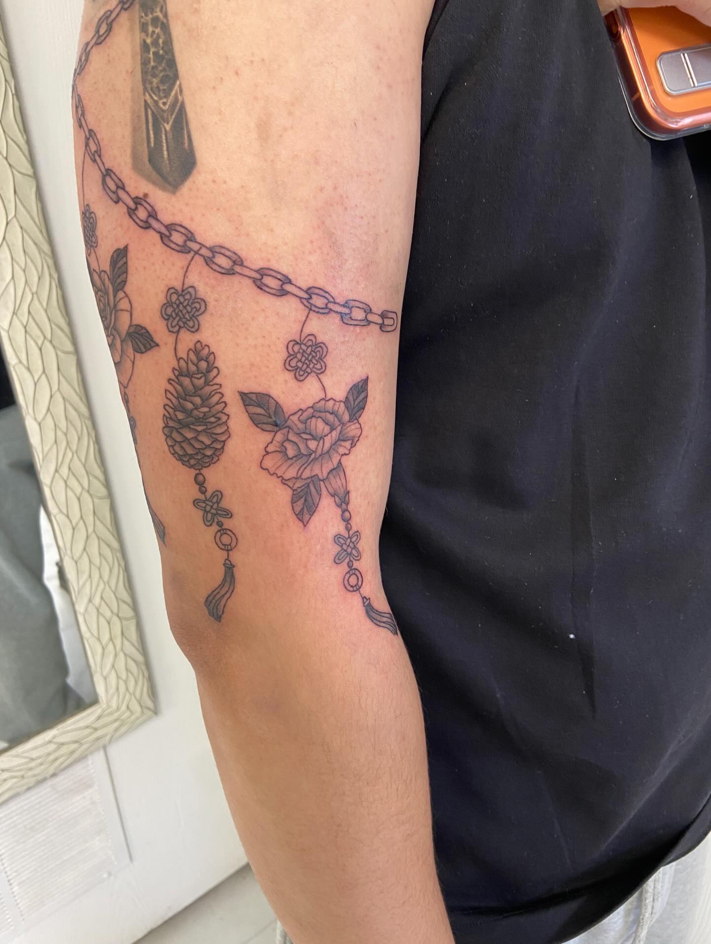 Chain with floral and pinecone motifs black and grey tattoo on the forearm by Sophia