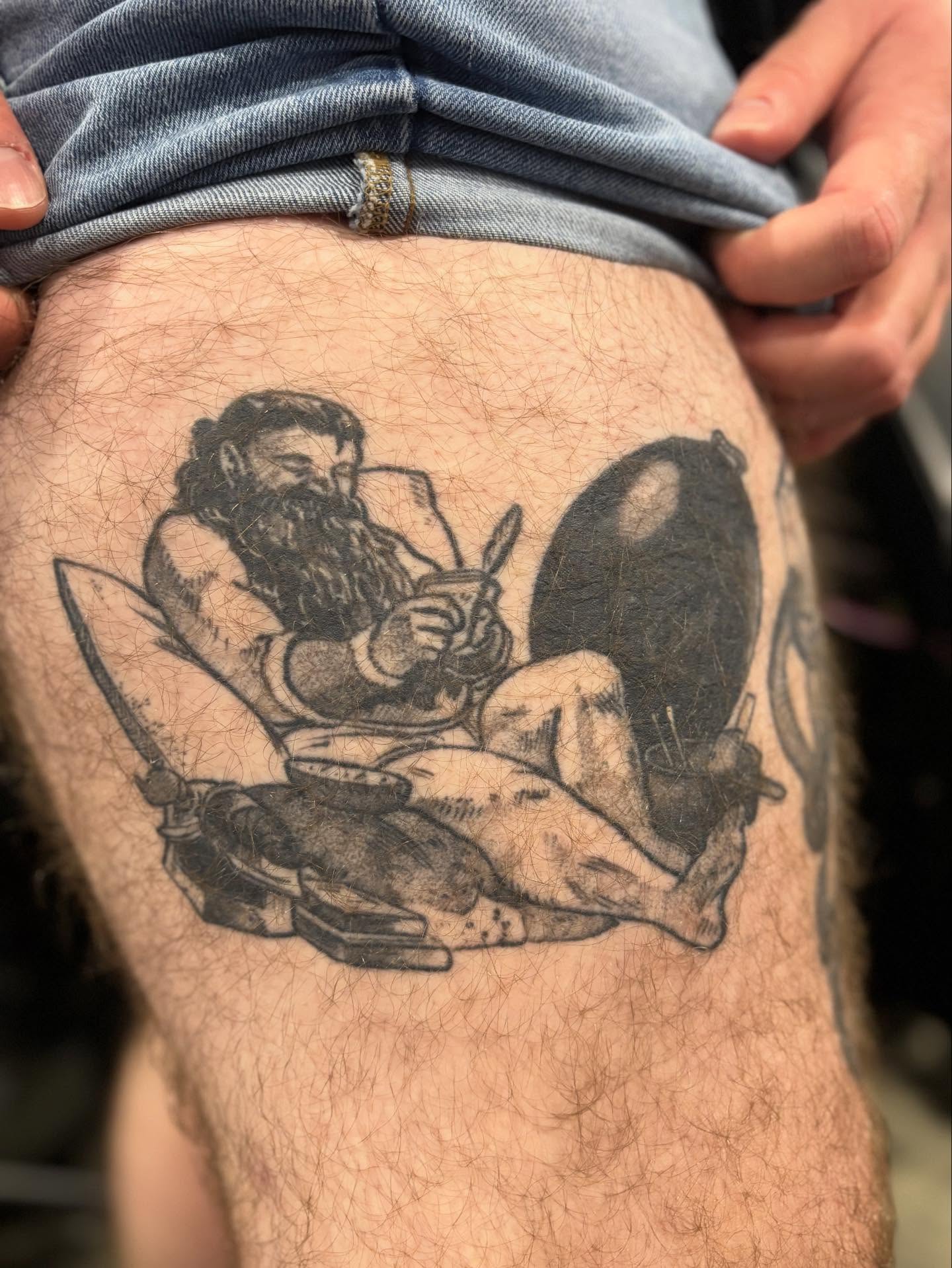 Bearded man reading black and grey tattoo on the thigh by Nick Tate