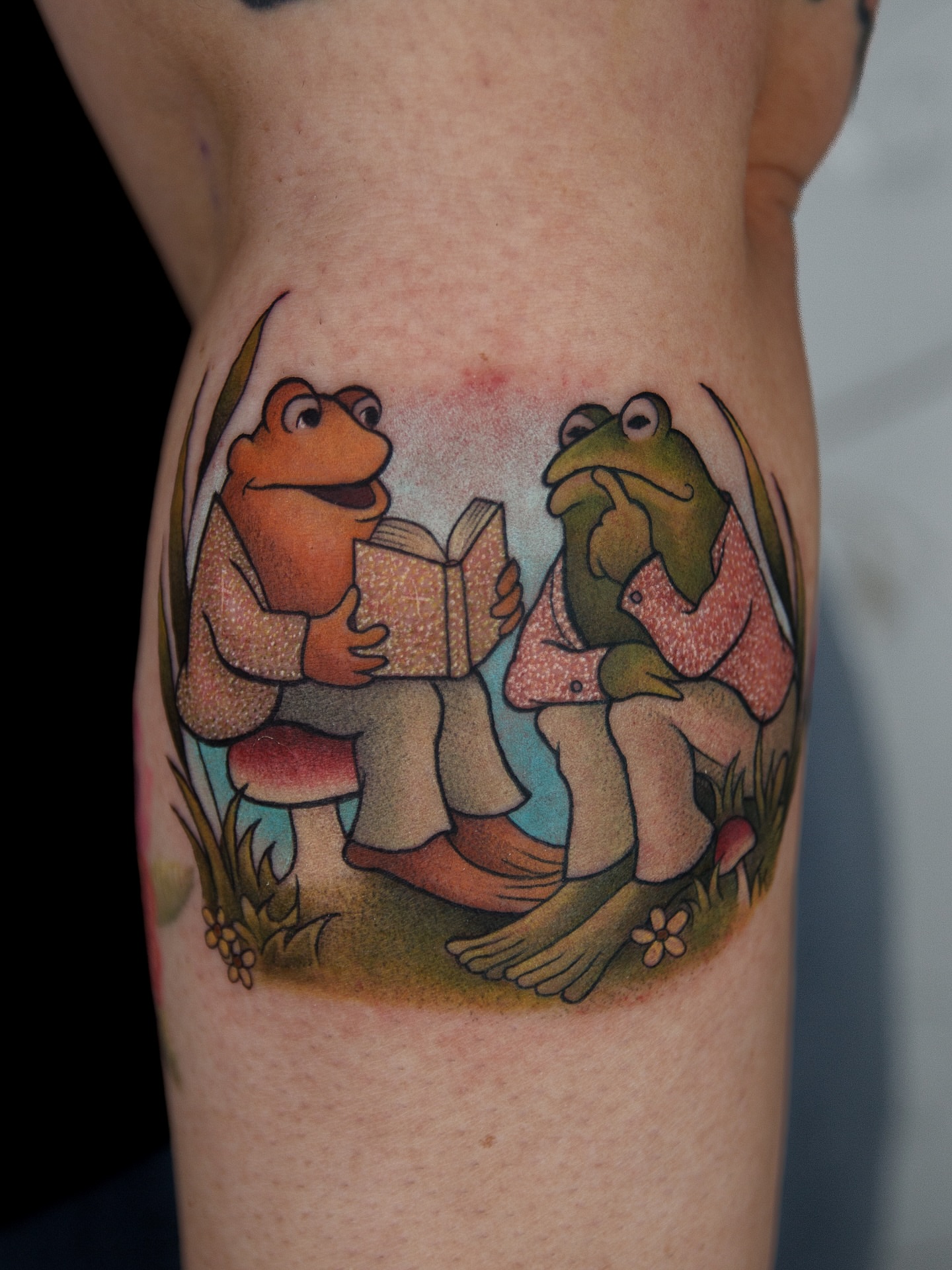 Frogs reading realism tattoo on the thigh by caliope yiannos