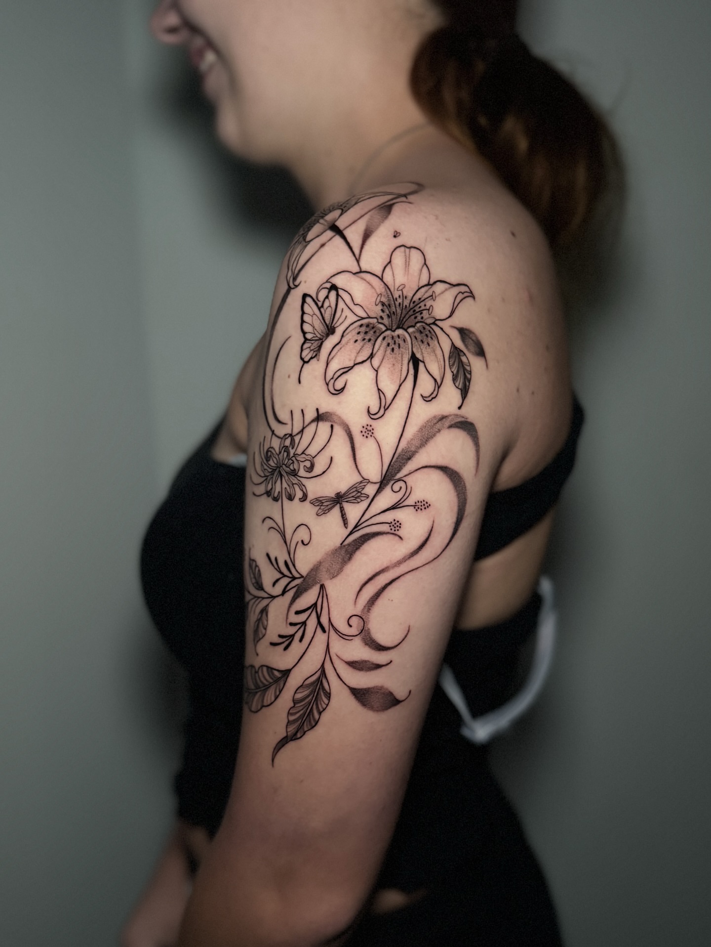 Lily and dragonfly black and grey tattoo on the upper arm by 𝕹𝖆𝖙