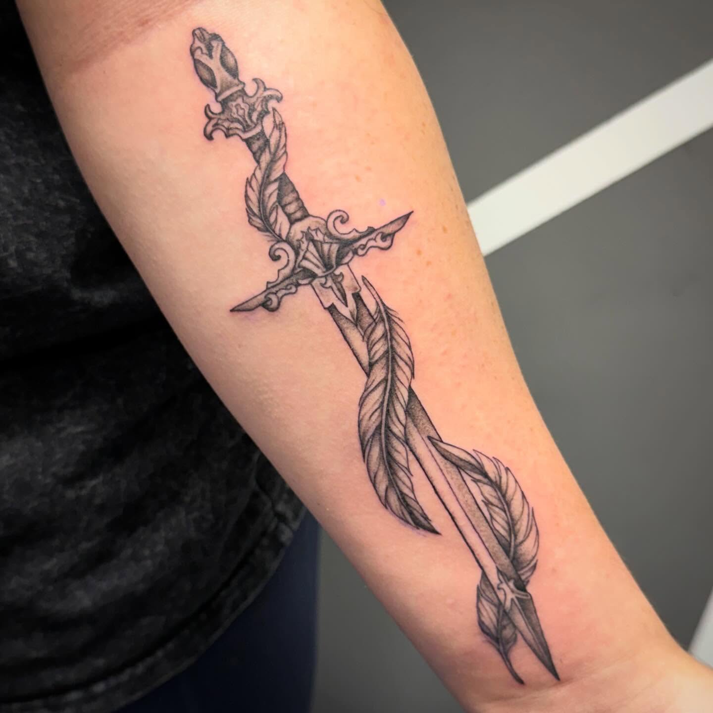 Sword and feathers black and grey tattoo on the forearm by Katie Rojas