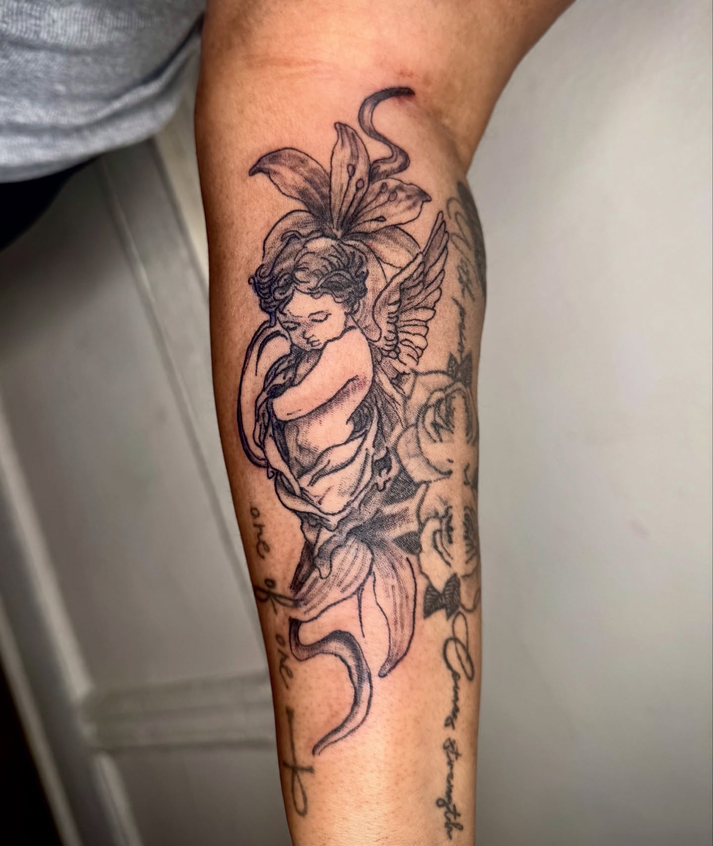 Cherub with lily black and grey tattoo on the forearm by JayyeSosa