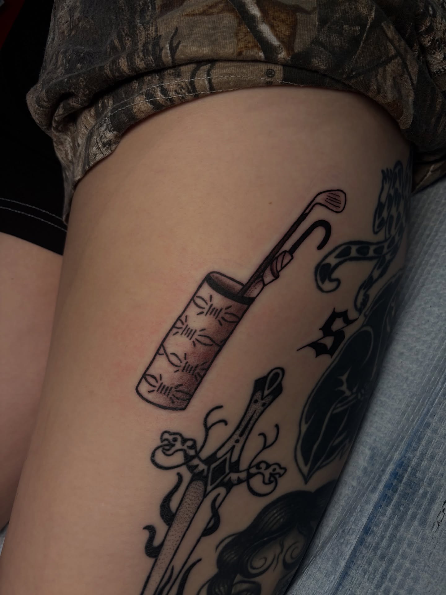 Golf club and drink black and grey tattoo on the thigh by Rachel