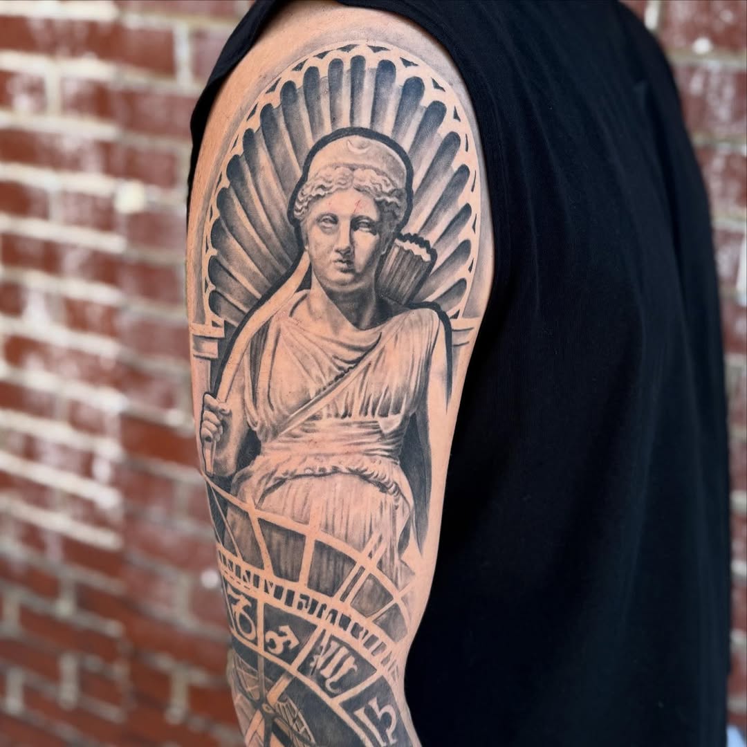 Statue of Artemis with astrological symbols realism tattoo on the upper arm by Jeremiah Warner
