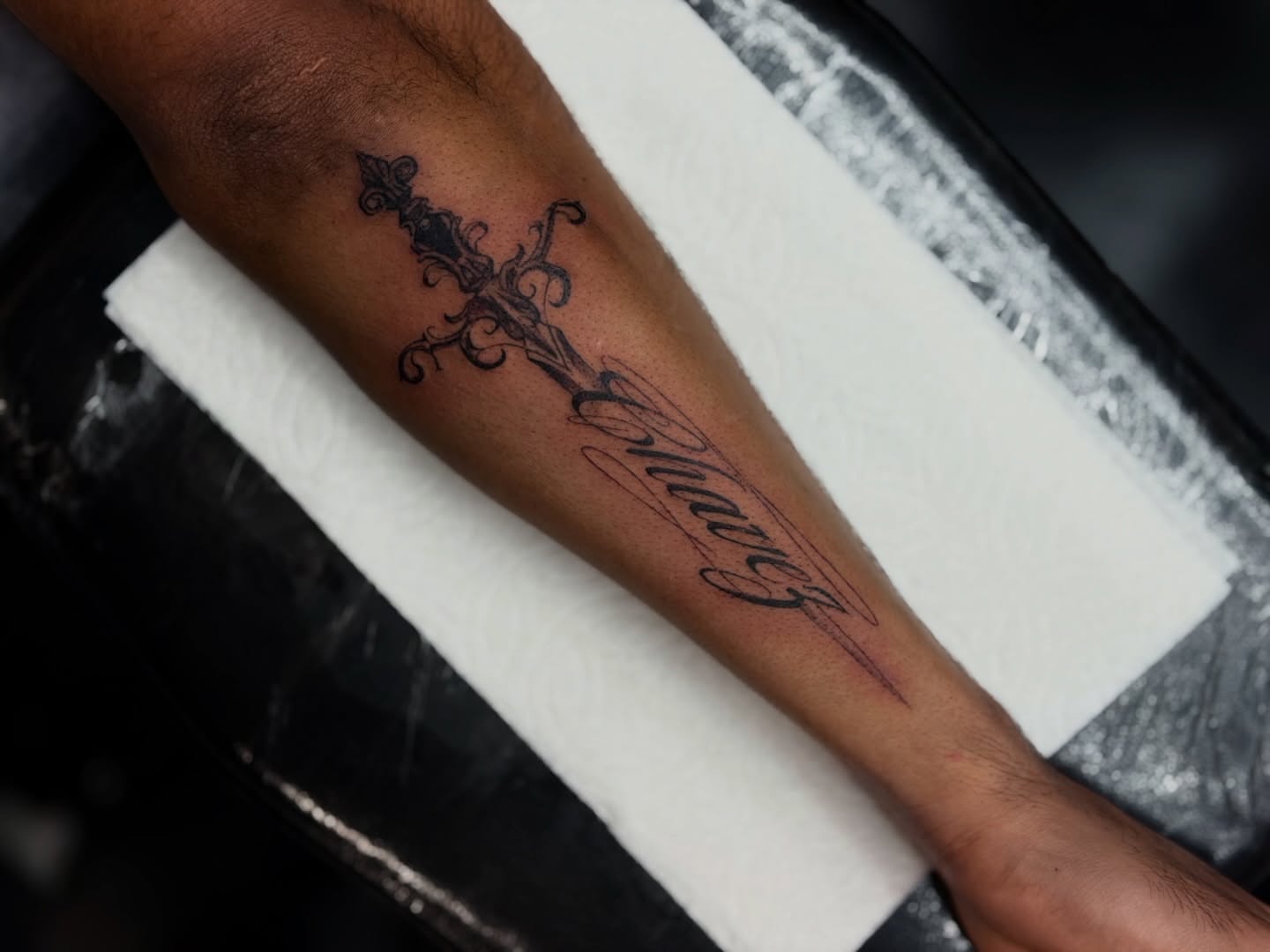 Sword and cursive text ornamental tattoo on the forearm by Bryan Nguyen