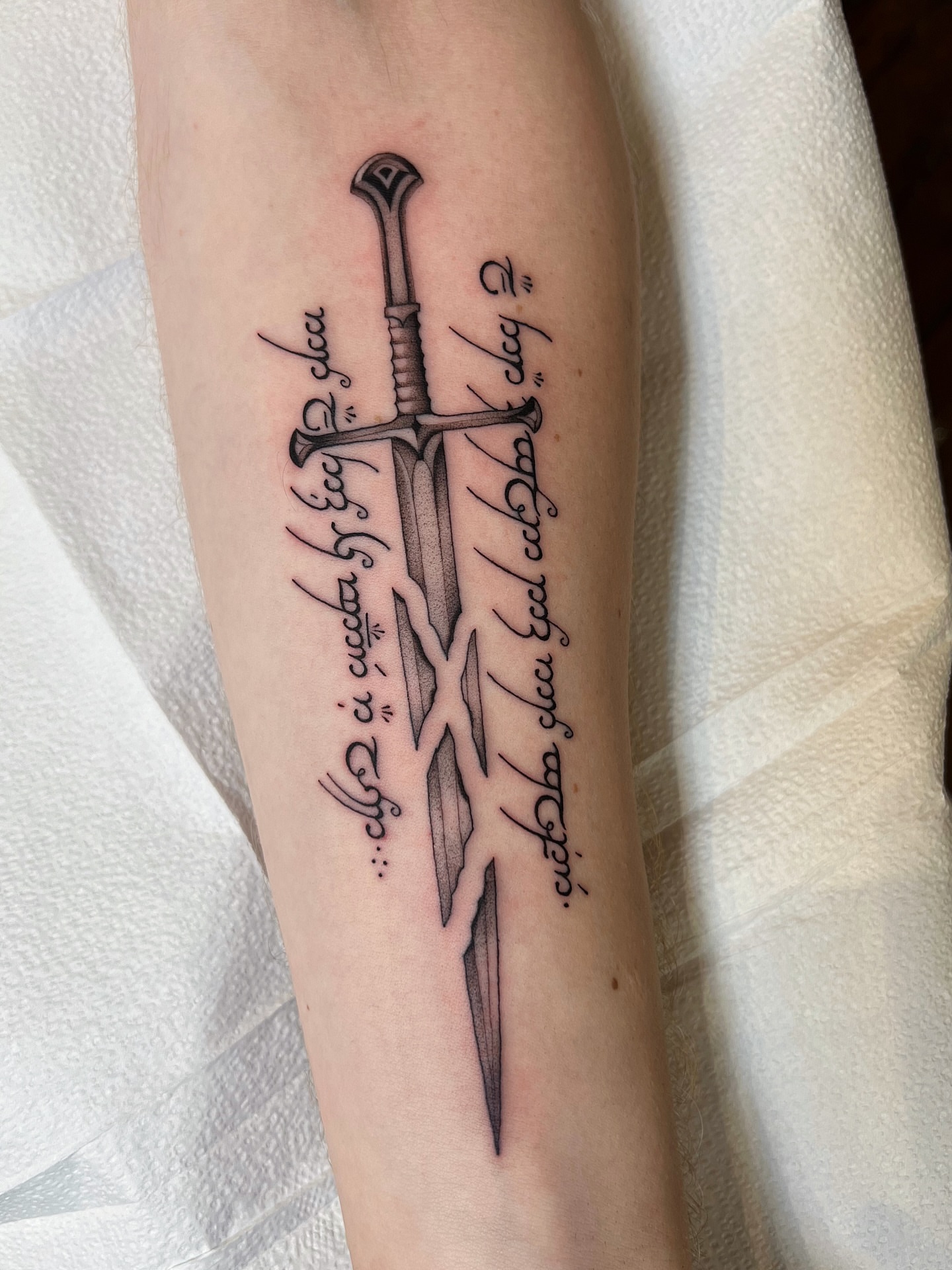 Sword and script black and grey tattoo on the forearm by Mickey