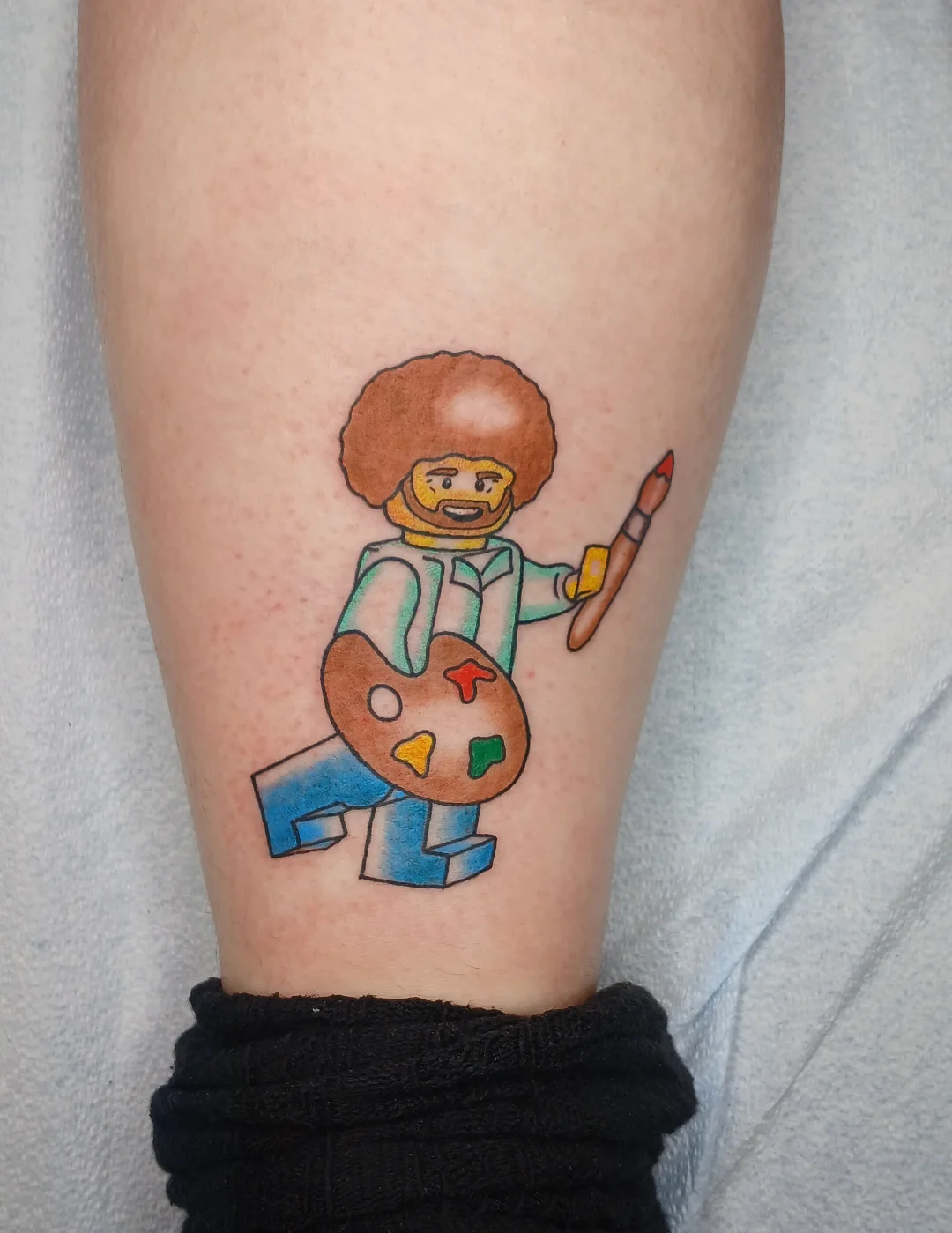 Bob Ross Lego figure illustrative tattoo on the forearm by Amber Tattoos
