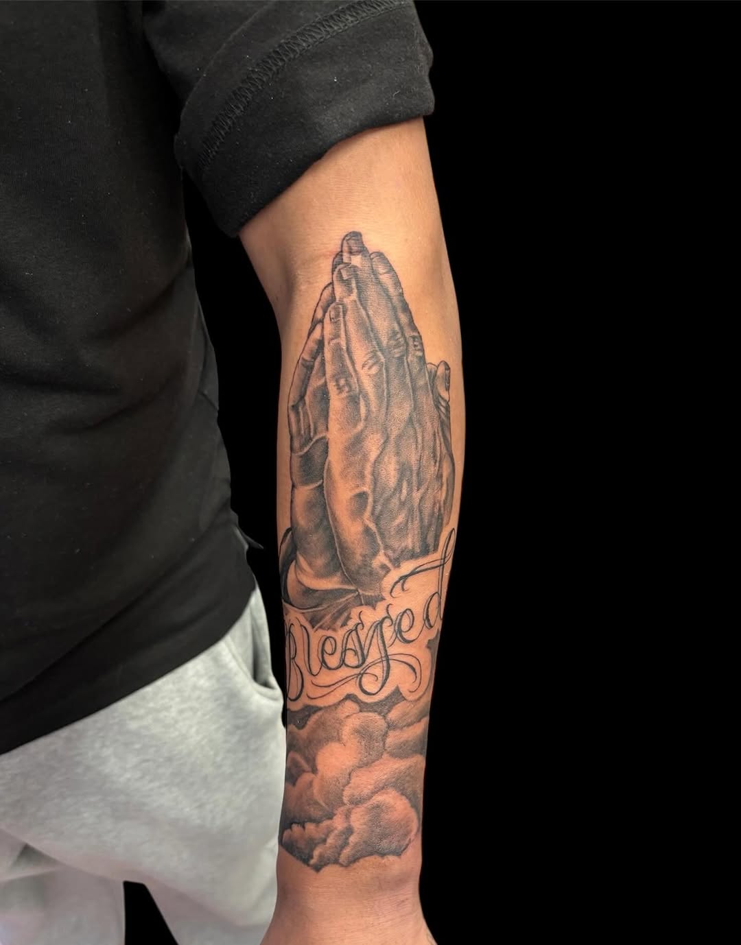 Praying hands with clouds and text Chicano tattoo on the forearm by @inkwitatlas