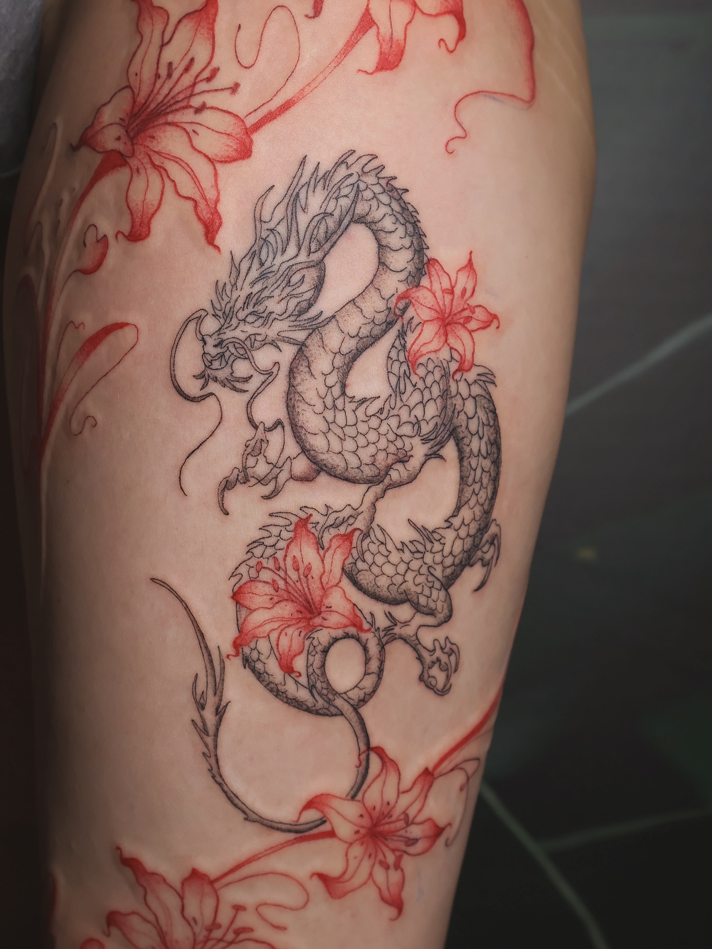 Dragon and flowers black and grey tattoo on the upper arm by 𝑭𝒊𝒏𝒆 𝑳𝒊𝒏𝒆 𝑻𝒂𝒕𝒕𝒐𝒐’𝒔