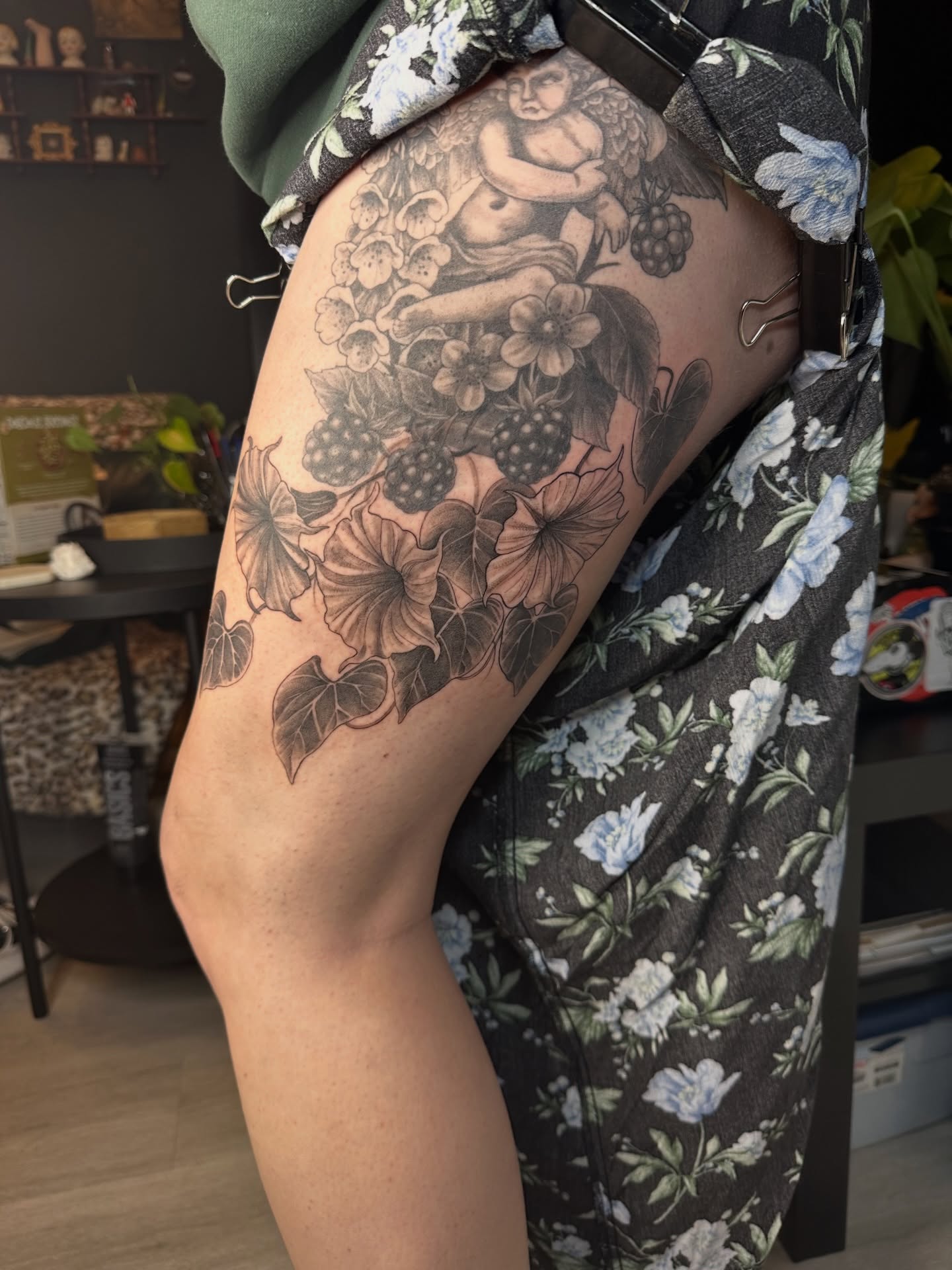 Cherub and floral bouquet black and grey tattoo on the thigh by Emma Thompson