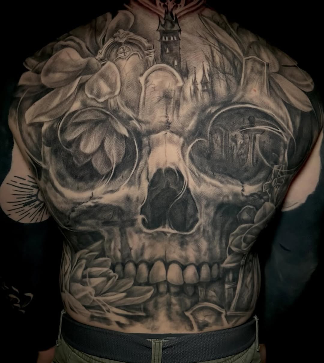 Skull with graveyard and flowers realism tattoo on the back by Comfy Chris Fettig