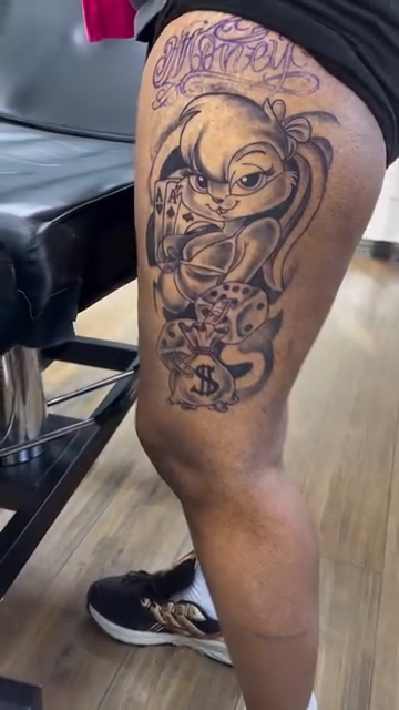 Cartoon character with dice and money lettering tattoo on the thigh by Inkman Parson