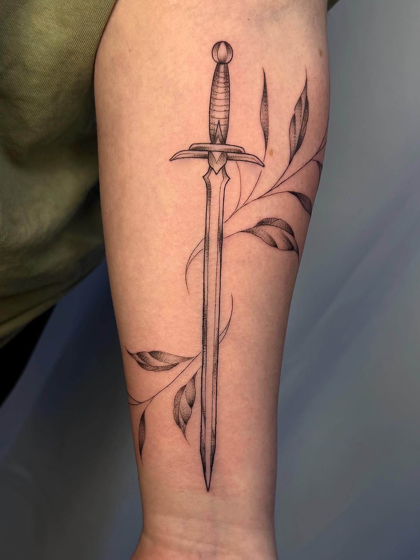 Sword and leaves black and grey tattoo on the forearm by sarah