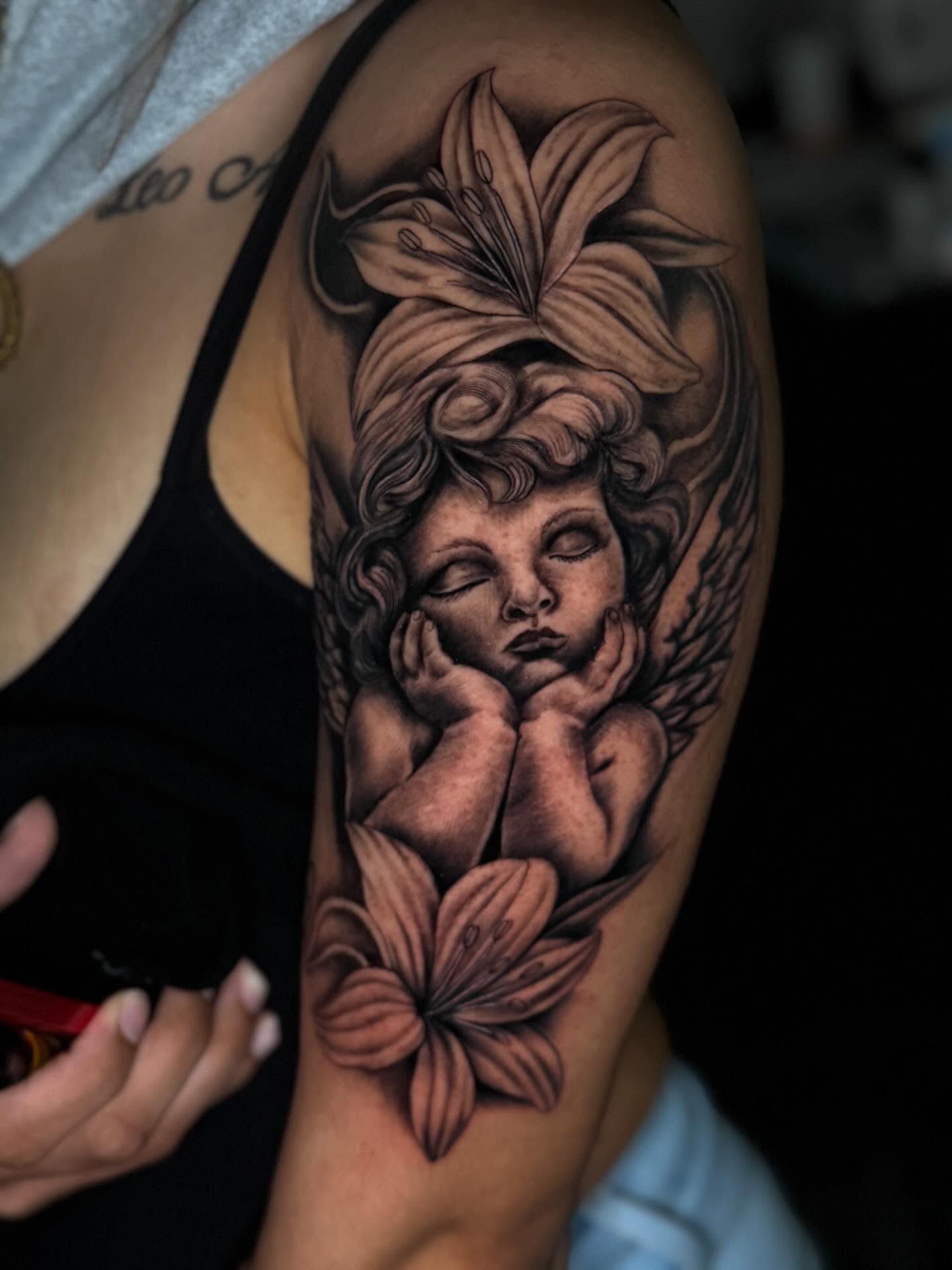 Cherub with lilies realism tattoo on the upper arm by Dany
