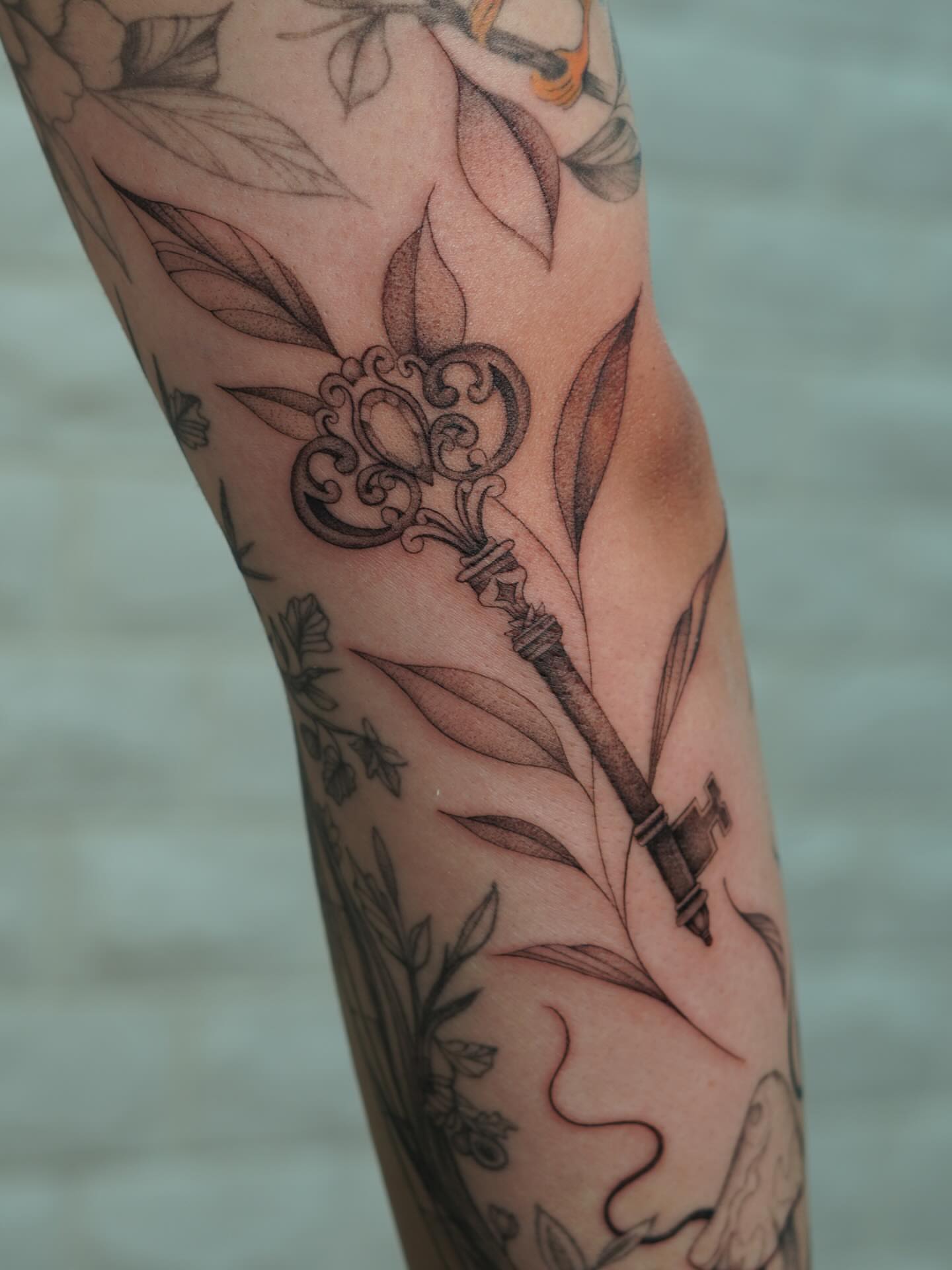 Key and leaves black and grey tattoo on the forearm by Danae Andino