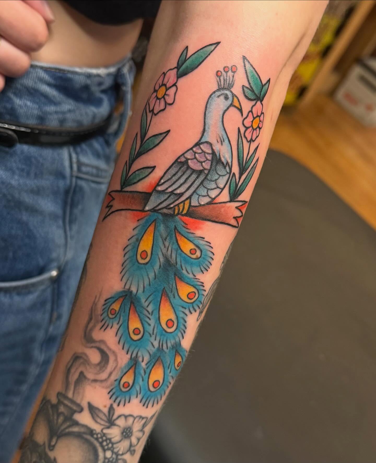 Peacock and floral wreath American traditional tattoo on the forearm by Madilynn Klassen