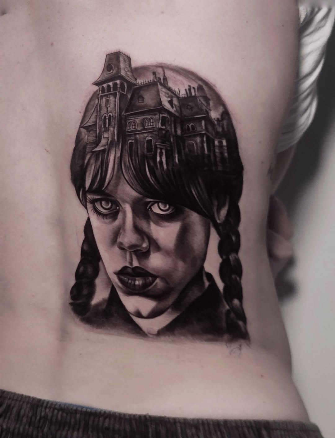 Wednesday Addams and haunted house realism tattoo on the ribs by Elidia Veliz