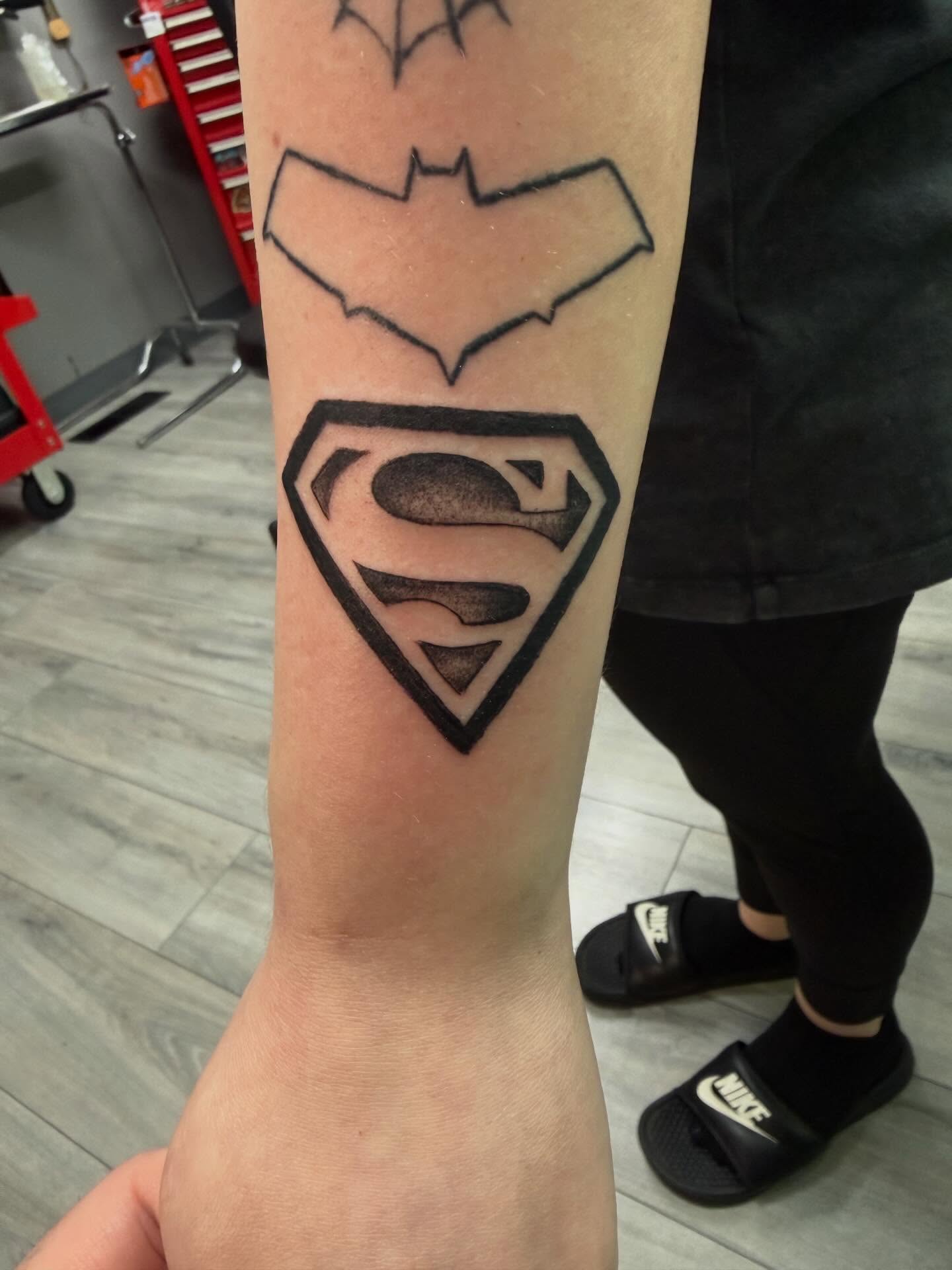 Batman and superman logos black and grey tattoo on the forearm by Daniel Smith